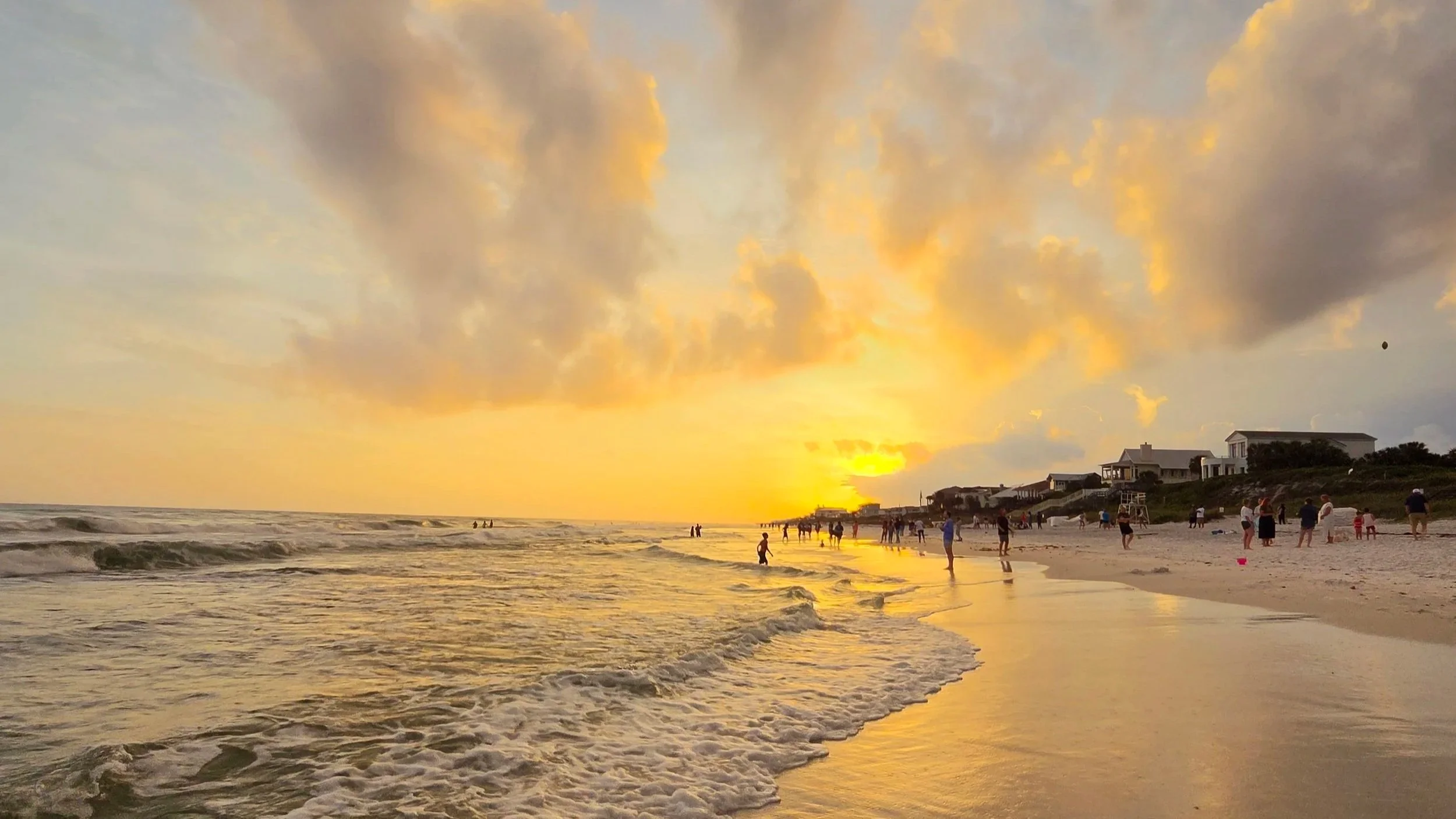 People enjoying a beach at sunset with waves, golden clouds, and beachfront homes