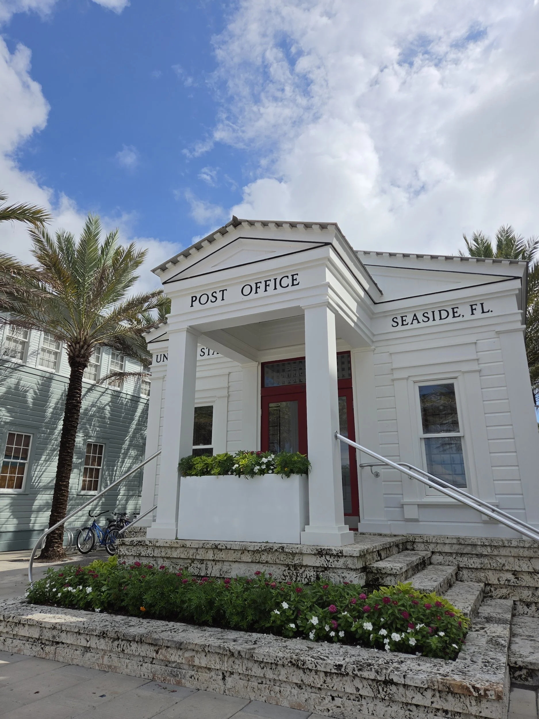 Historic white Post Office building labeled “Seaside, FL,” with palm trees and flower beds under a partly cloudy blue sky.