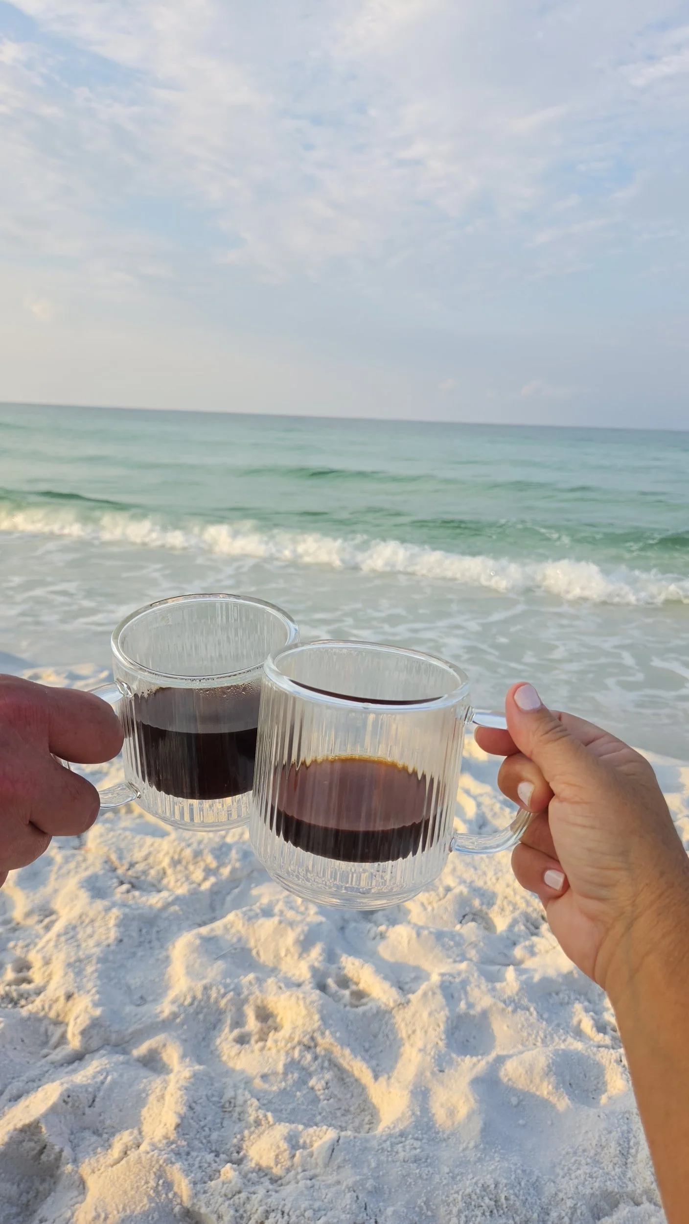 Two hands holding glass mugs of coffee on a white sandy beach with turquoise ocean waves