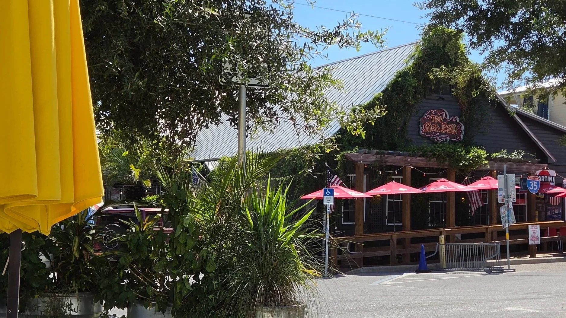 Outdoor restaurant and bar patio with red umbrellas, tropical plants, and a neon-style “The Red Bar” sign.
