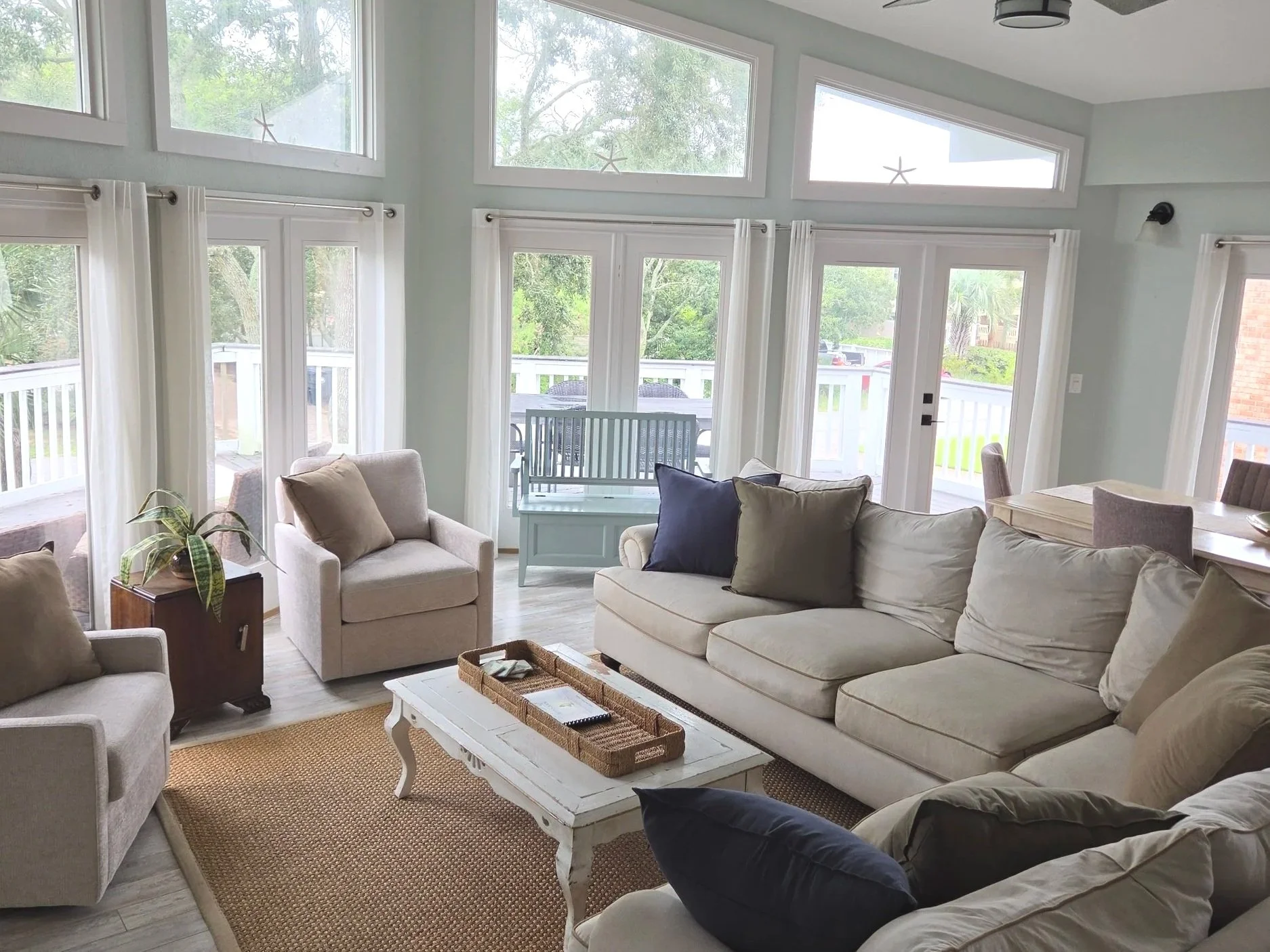 Bright coastal-style living room with white sofas, armchairs, woven rug, coffee table, large windows, and deck views