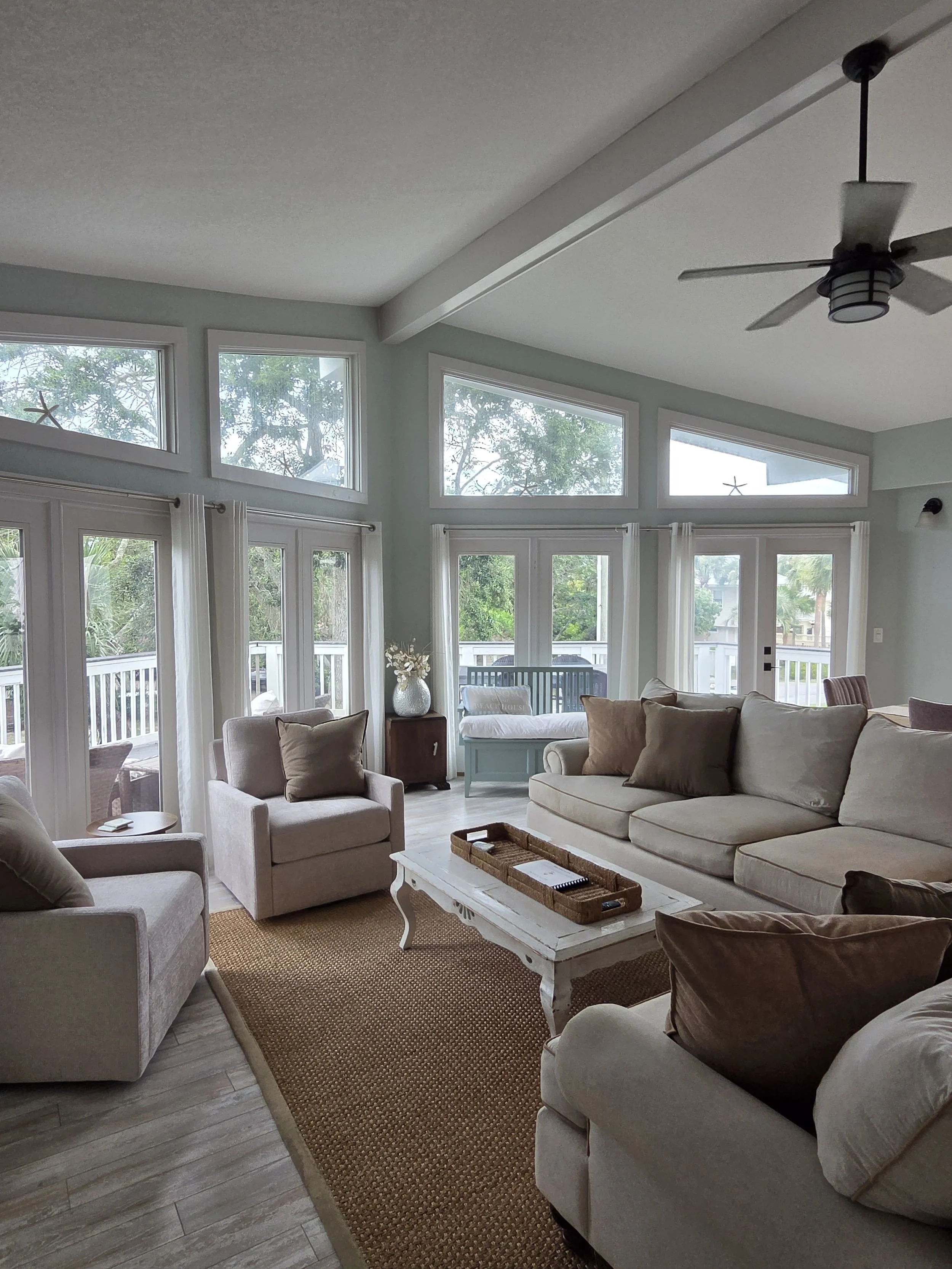 Bright coastal-style living room with white sofas, armchairs, woven rug, coffee table, ceiling fan, and large windows overlooking greenery.