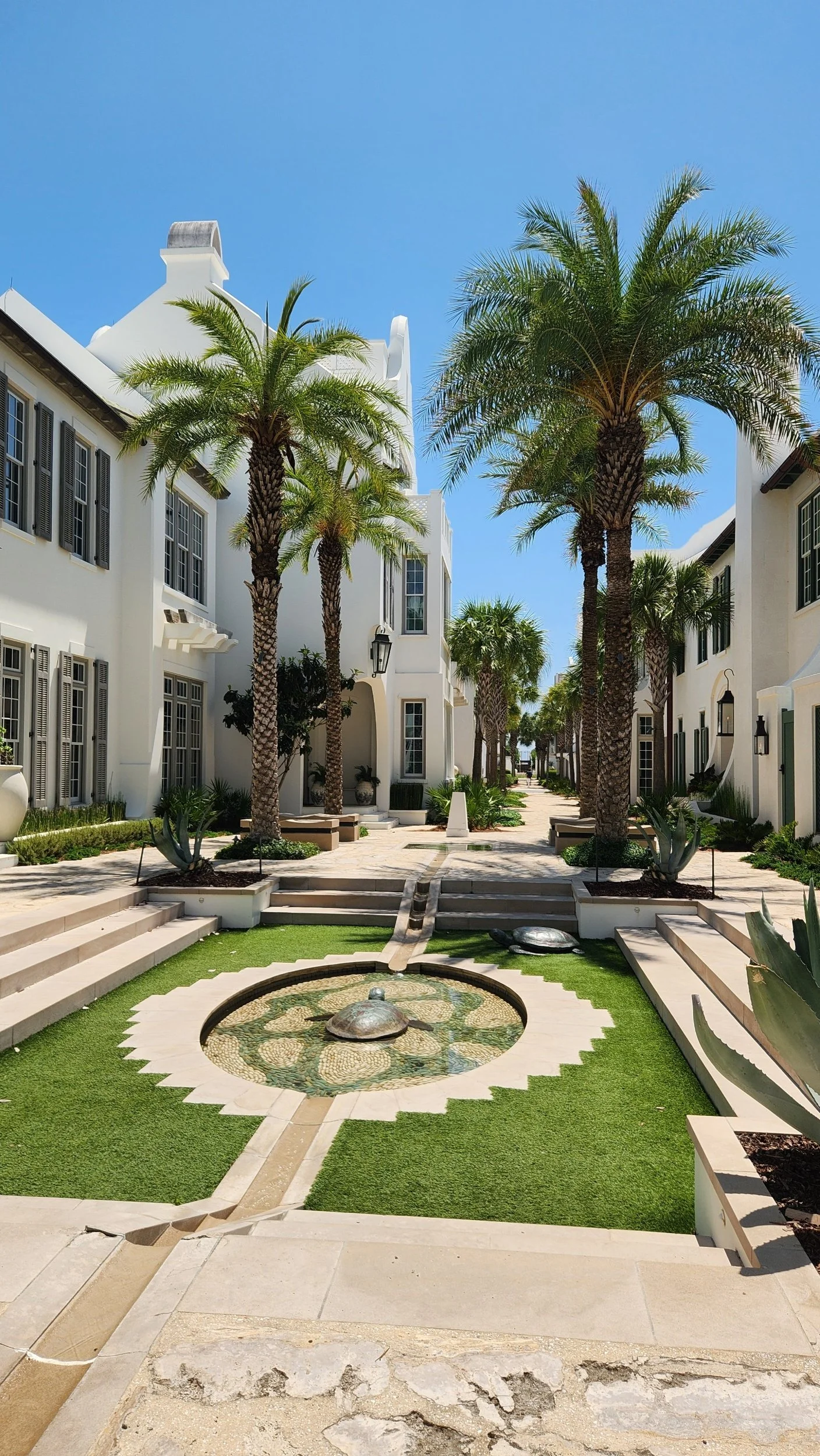 Palm-lined courtyard with white Mediterranean-style buildings, tiled fountain, landscaped pathways, and bright blue sky