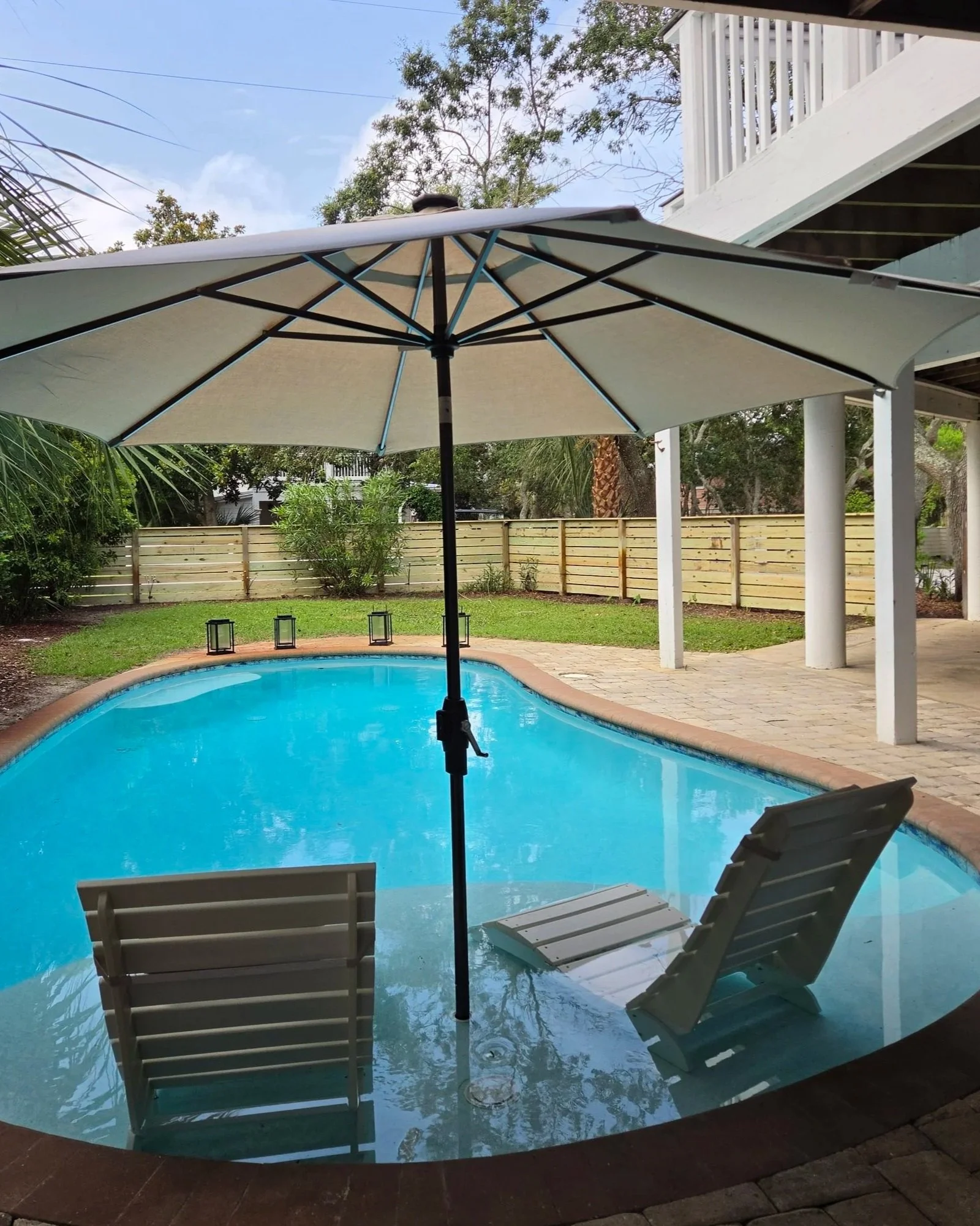Backyard swimming pool with lounge chairs beneath a large patio umbrella, surrounded by tropical landscaping and a covered porch.
