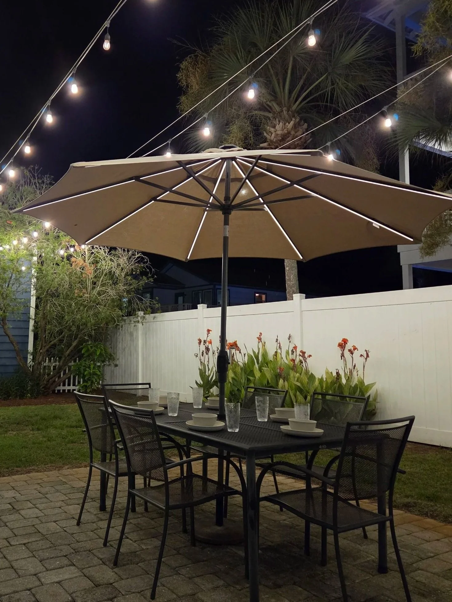 Outdoor patio dining area at night with a large umbrella, set table, string lights, tropical plants, and black metal chairs.