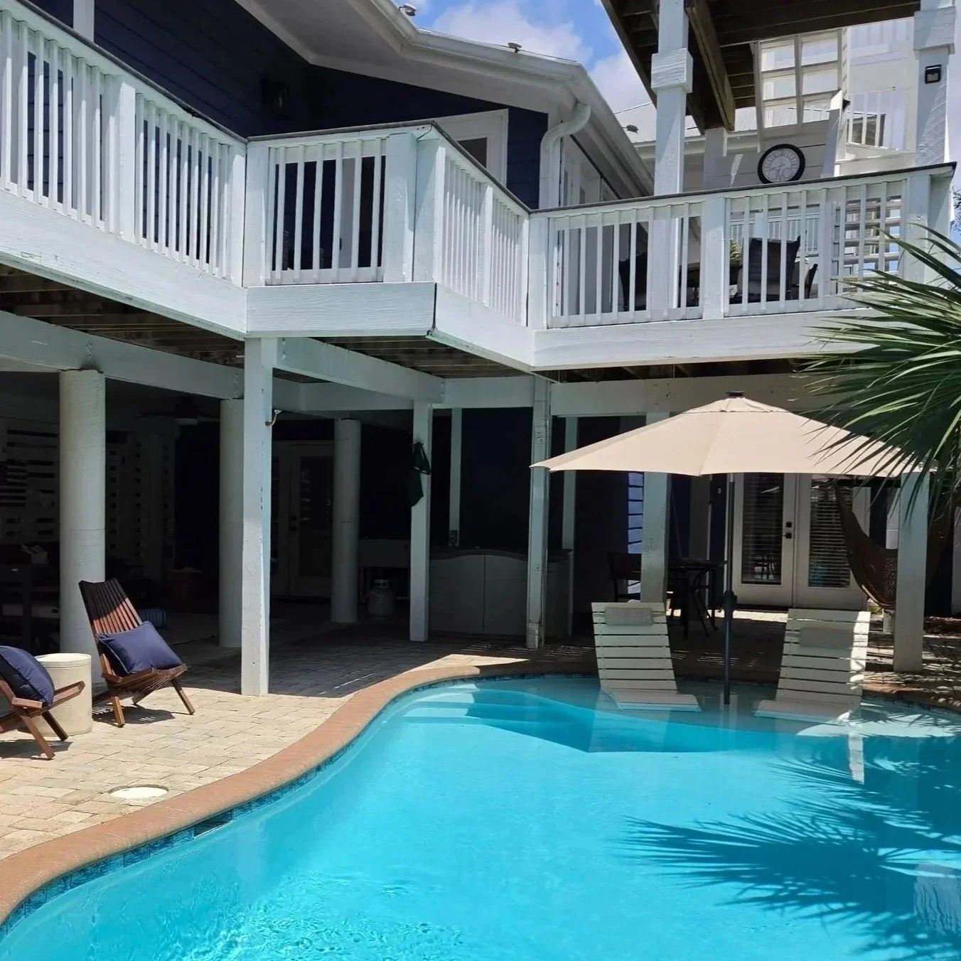 Backyard swimming pool beside a white-railed coastal house with lounge chairs, patio umbrella, and palm tree