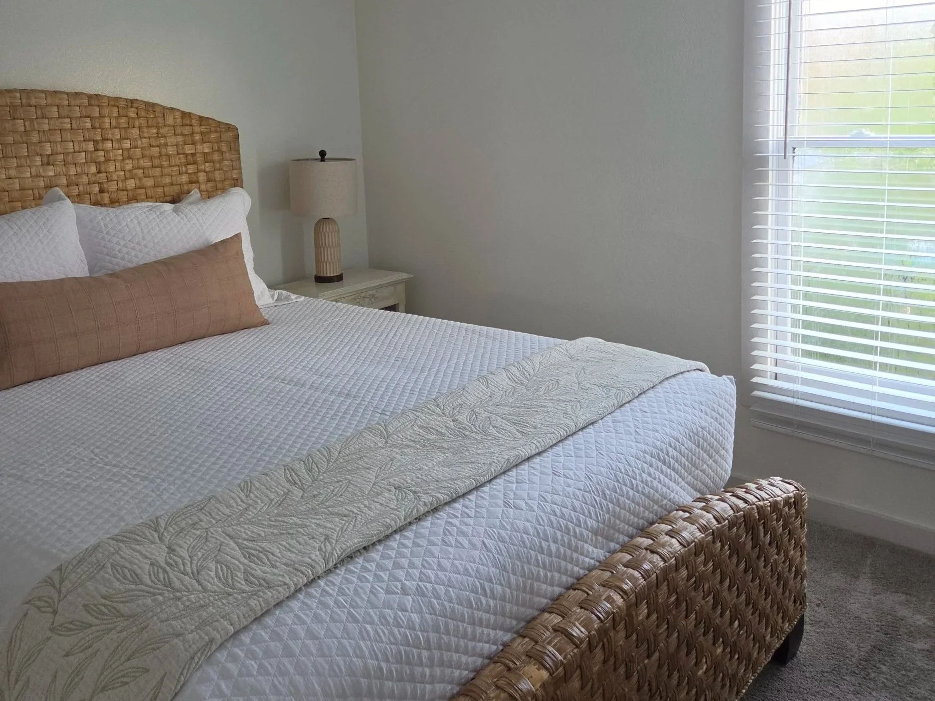 Bright bedroom with a woven wicker bed, white quilted bedding, neutral pillows, bedside lamp, and window blinds