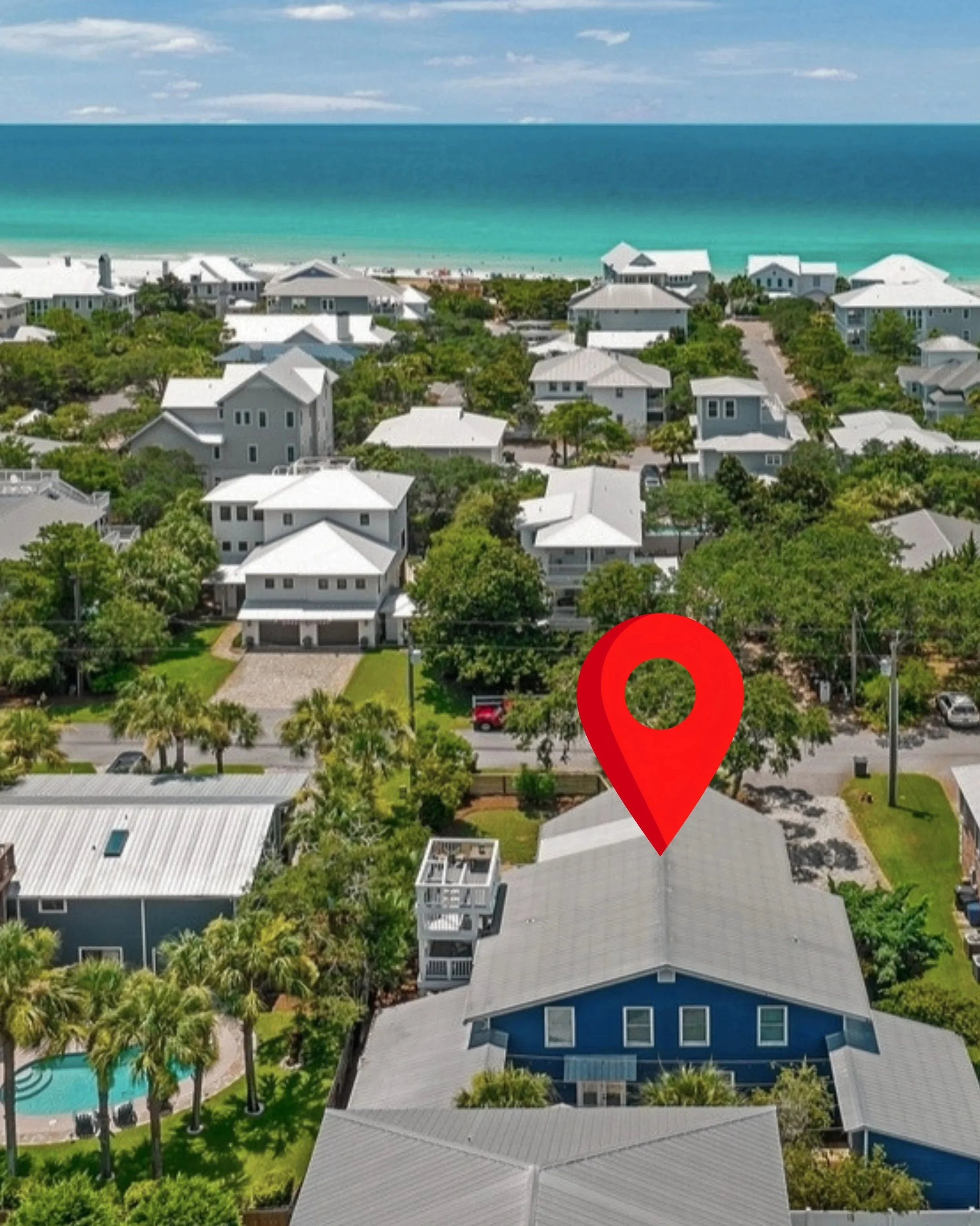 Aerial view of tropical coastal homes, palm trees, turquoise ocean, and a large red map pin marking a property