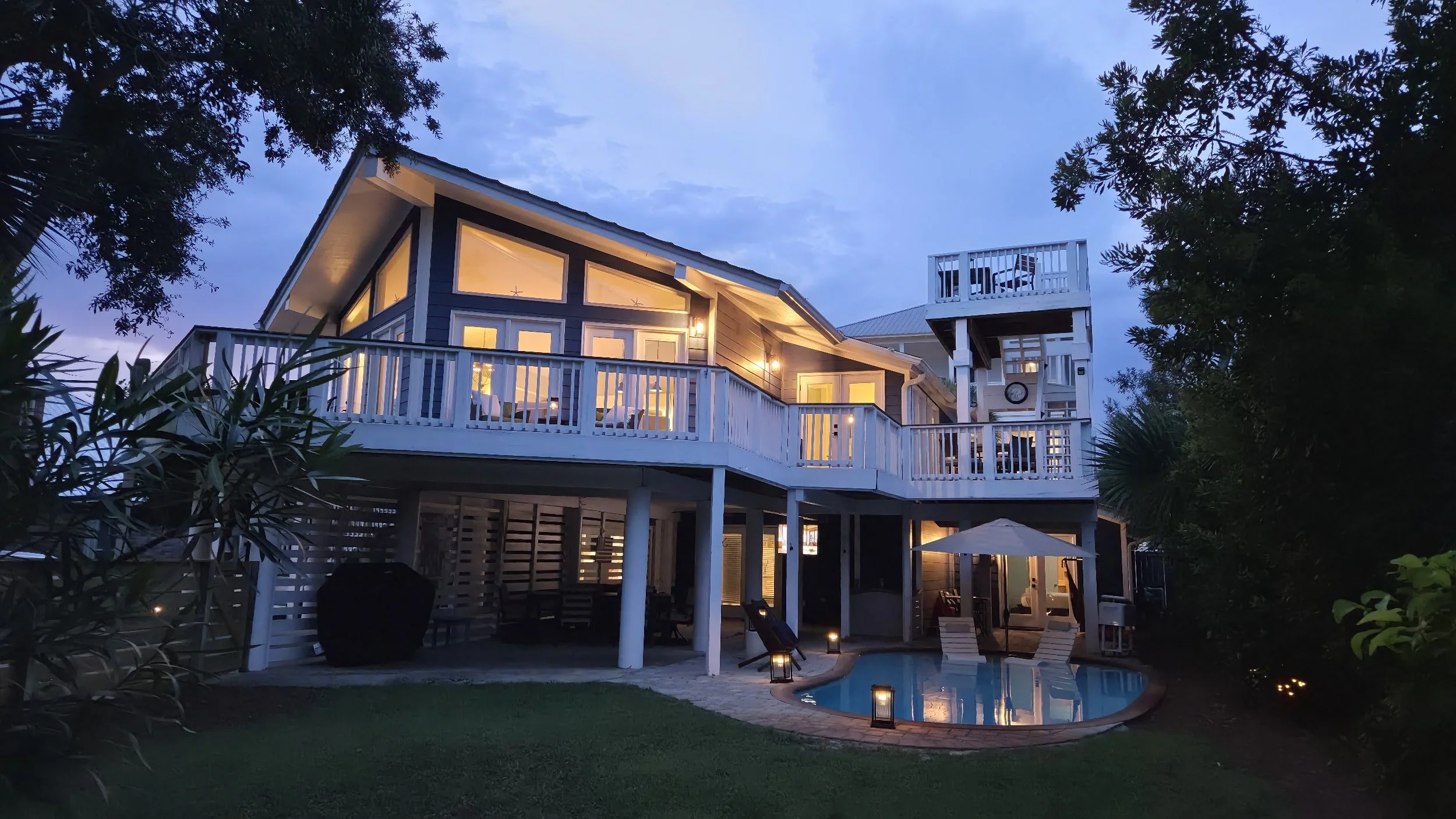 Large illuminated coastal-style home with wraparound decks, lush tropical landscaping, and a backyard swimming pool at dusk