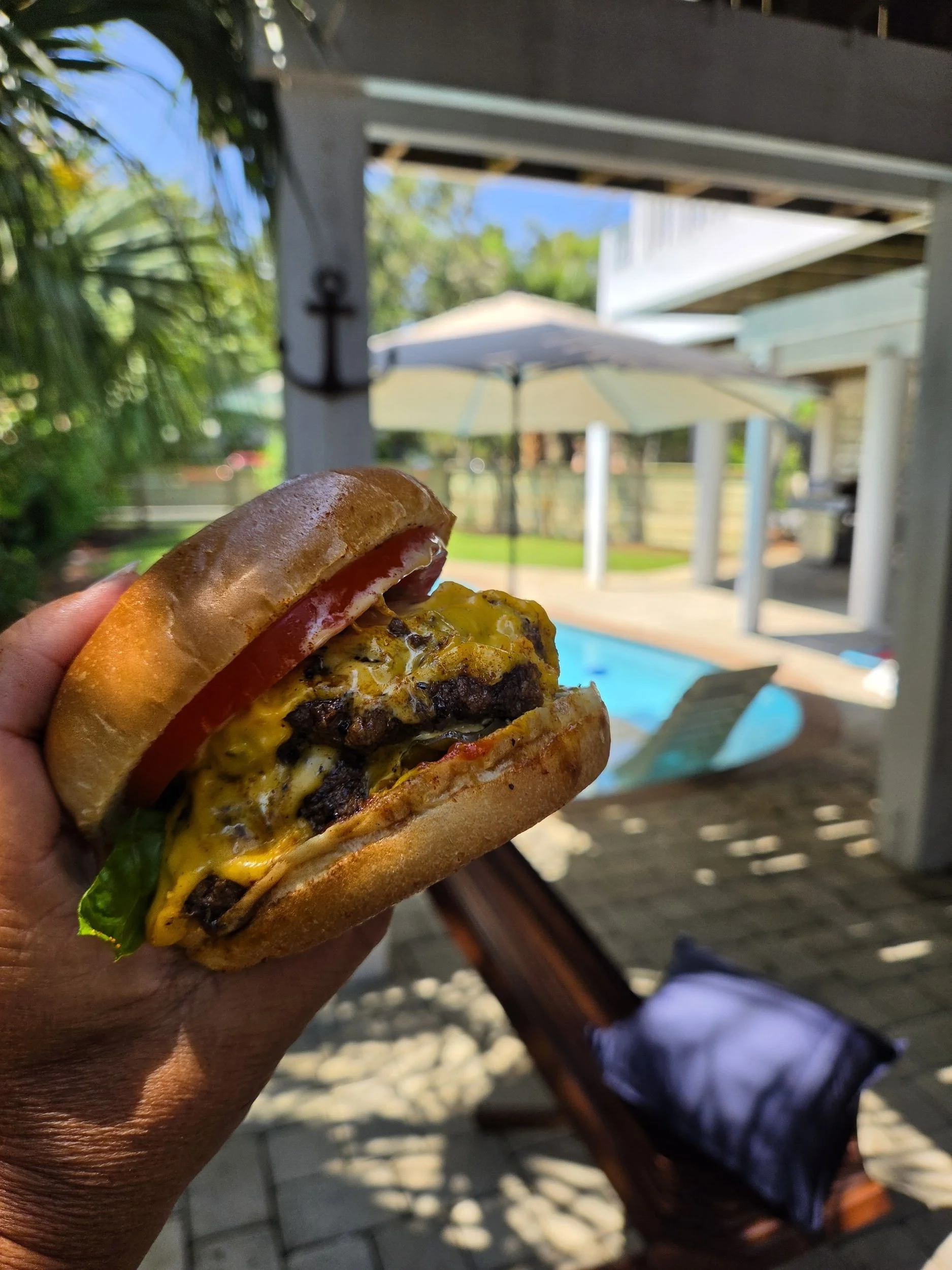 Hand holding a double cheeseburger with tomato and lettuce outdoors beside a swimming pool
