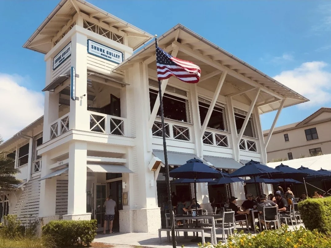 Shuck Gulley Oyster Bar restaurant with outdoor patio seating, blue umbrellas, and an American flag