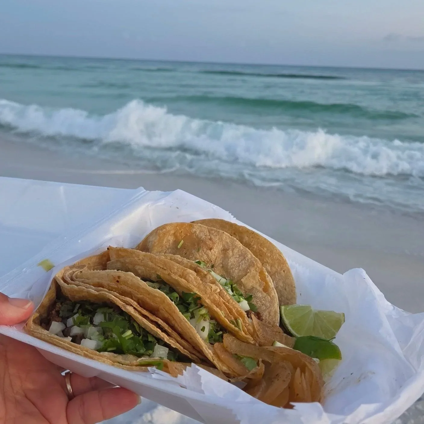 Tray of Mexican street tacos with cilantro, diced onions, and lime on a beach overlooking ocean waves