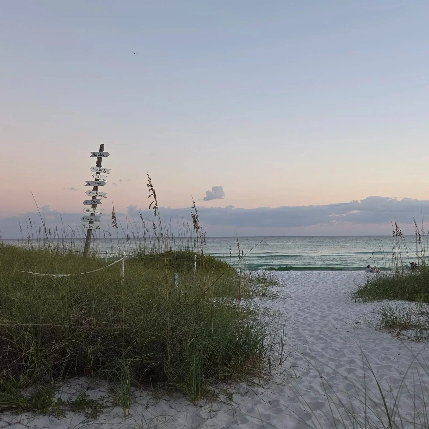 Sunset beach access through coastal dunes, with sea oats, ocean waves, and a signpost pointing to destinations including New York and Atlanta.