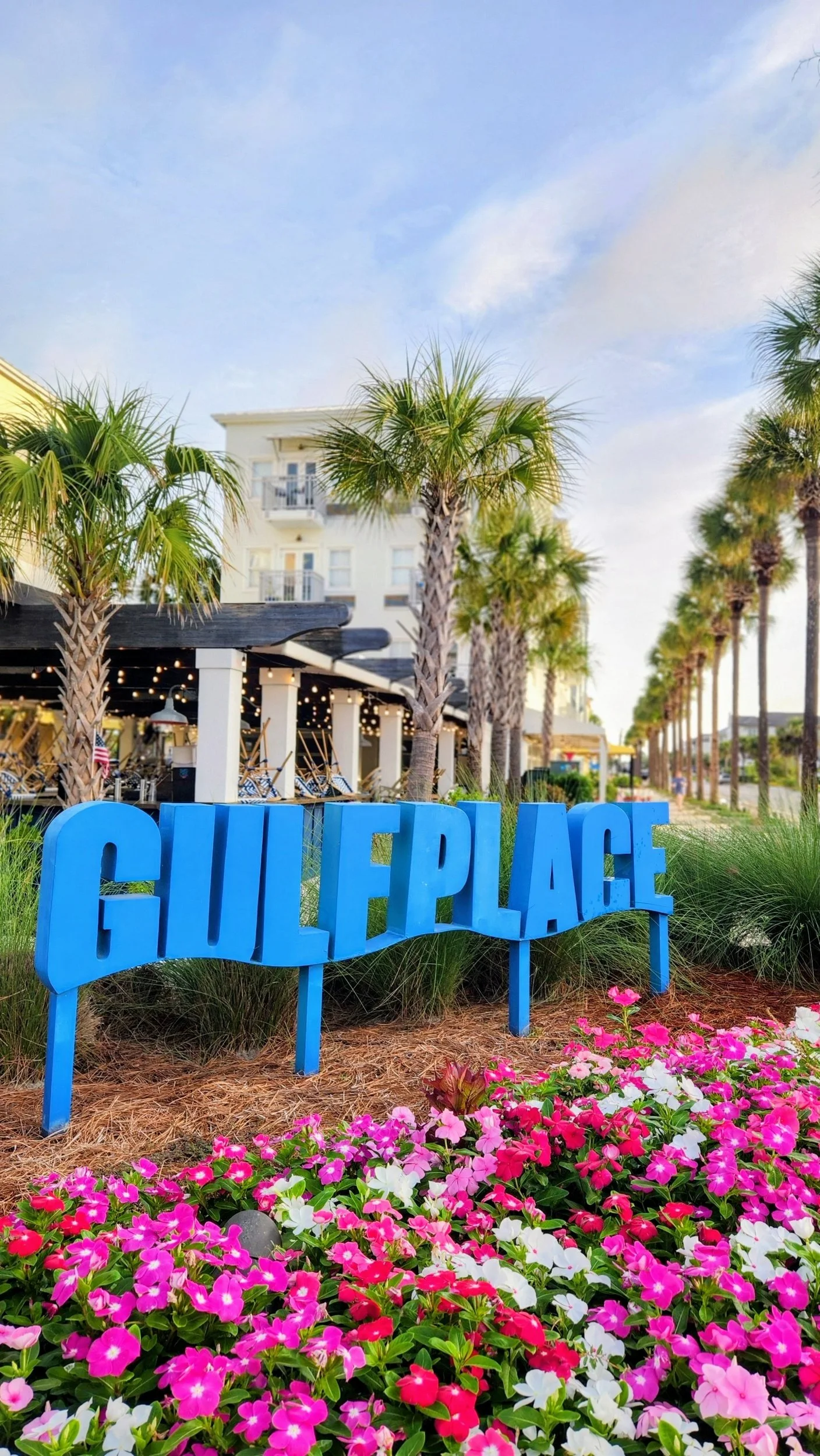 Large blue Gulf Place sign surrounded by palm trees and vibrant pink and white flowers outside a coastal resort.
