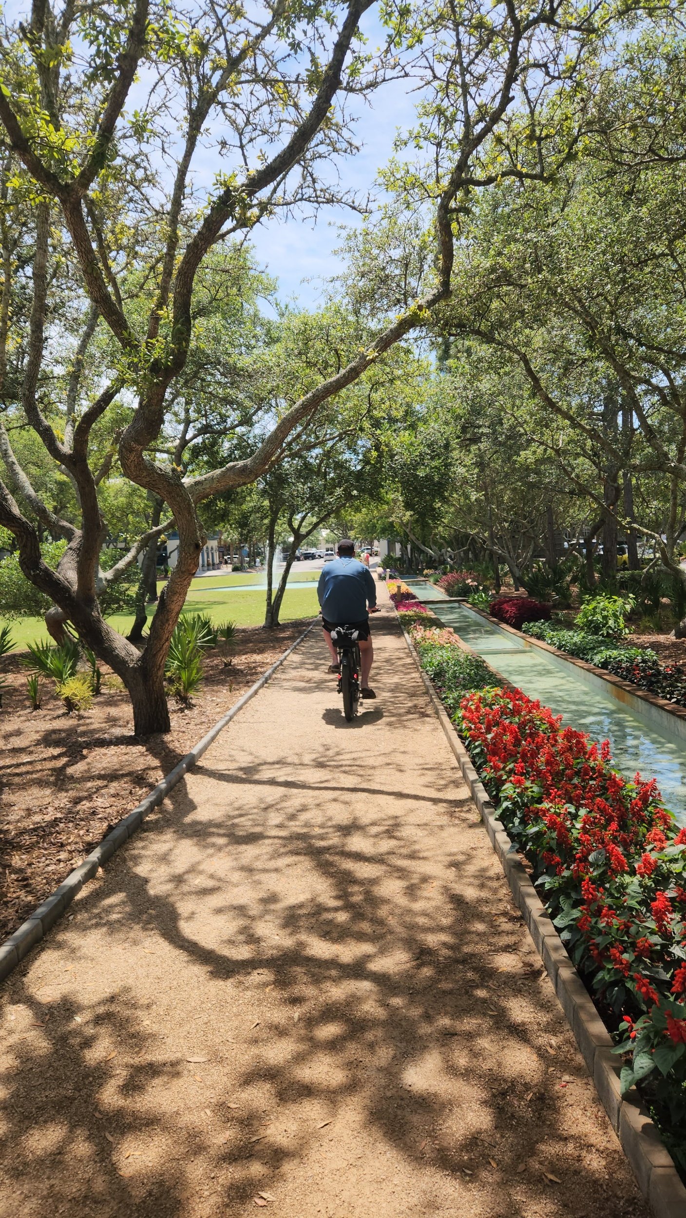 Person riding a bicycle along a tree-lined garden path beside a flower-bordered water channel.