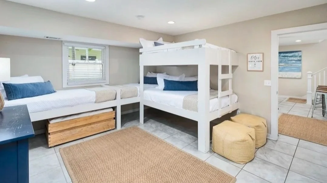 Bright beach-style bedroom with white bunk beds, twin bed, blue accent pillows, woven rug, and yellow ottomans
