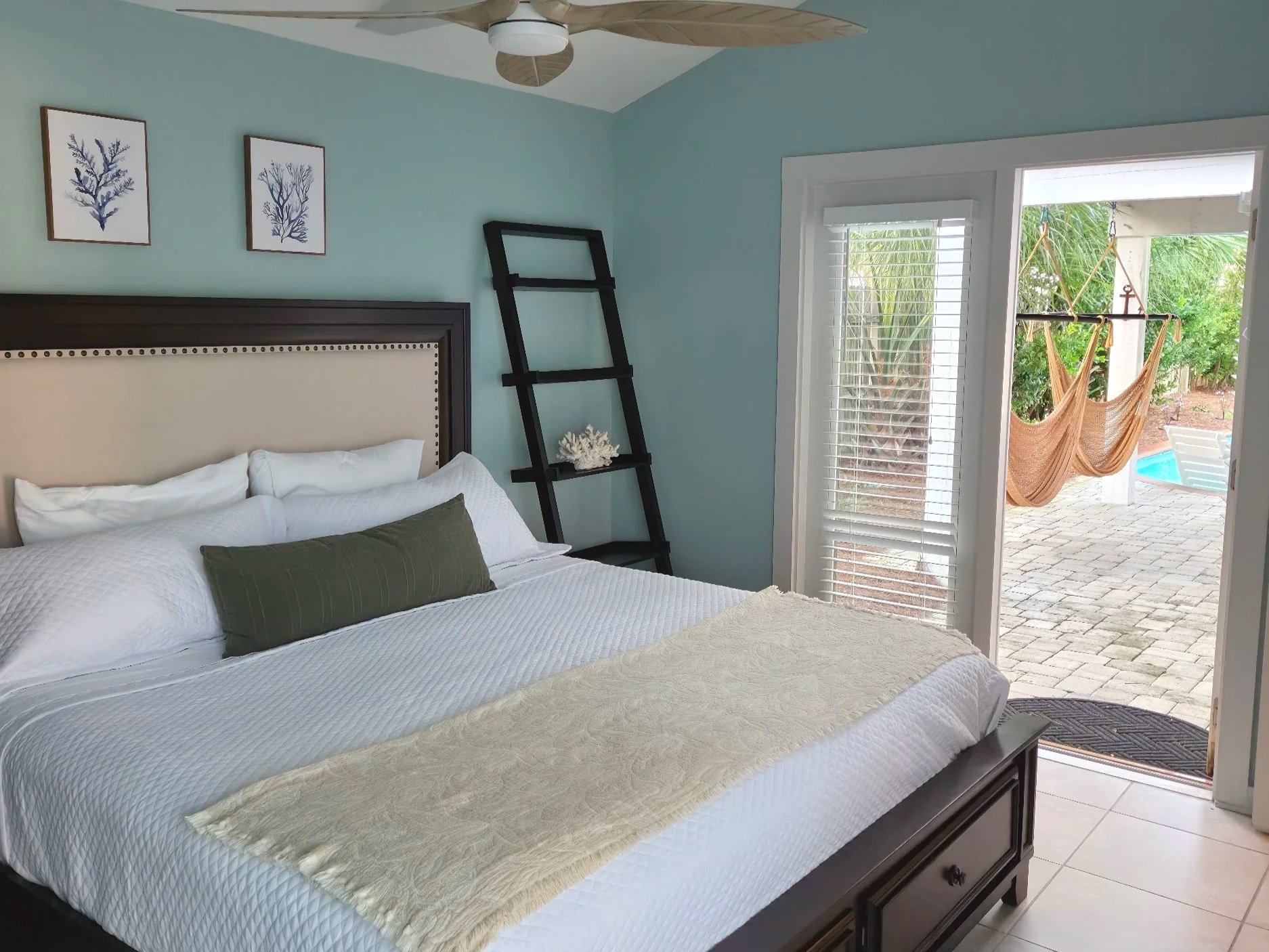 Bright coastal bedroom with a neatly made bed, blue walls, coral artwork, ceiling fan, ladder shelf, and open patio doors overlooking hammocks and a pool.