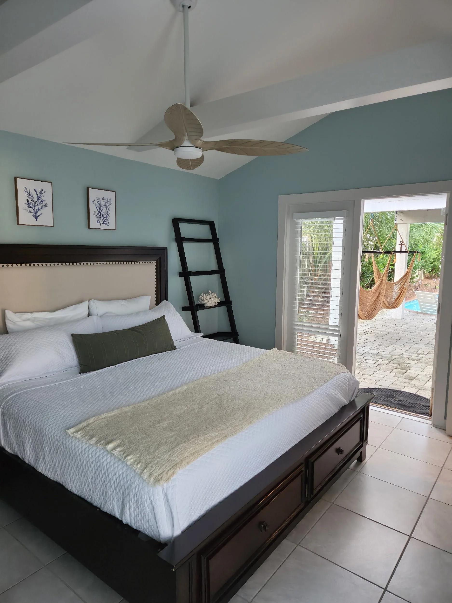 Bright coastal-style bedroom with a neatly made bed, wooden ceiling fan, blue walls, framed botanical art, and open doors leading to a patio with a hammock and pool.