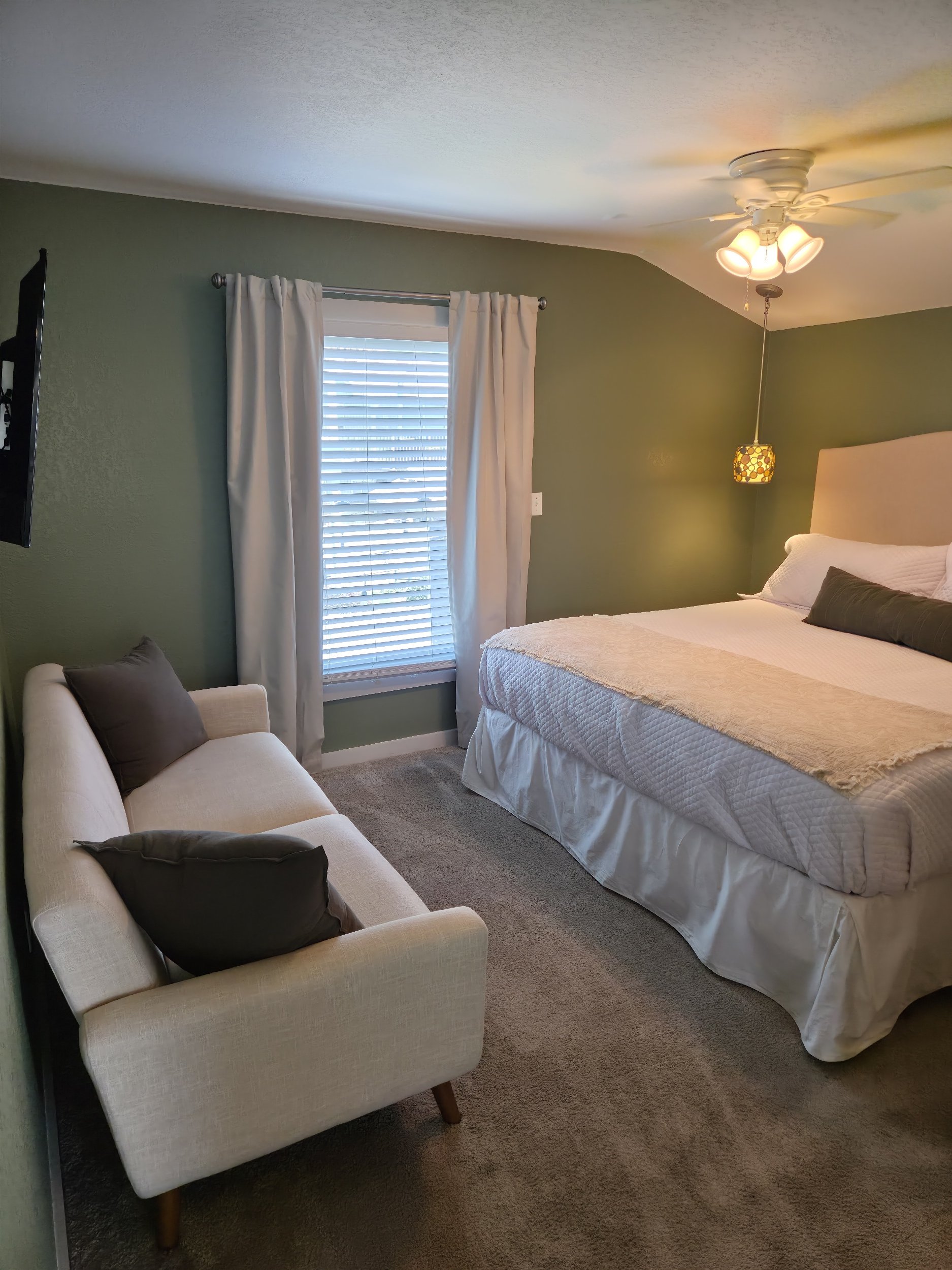 Cozy bedroom with a white bed, green walls, window with white curtains and blinds, armchair, and ceiling fan with lights