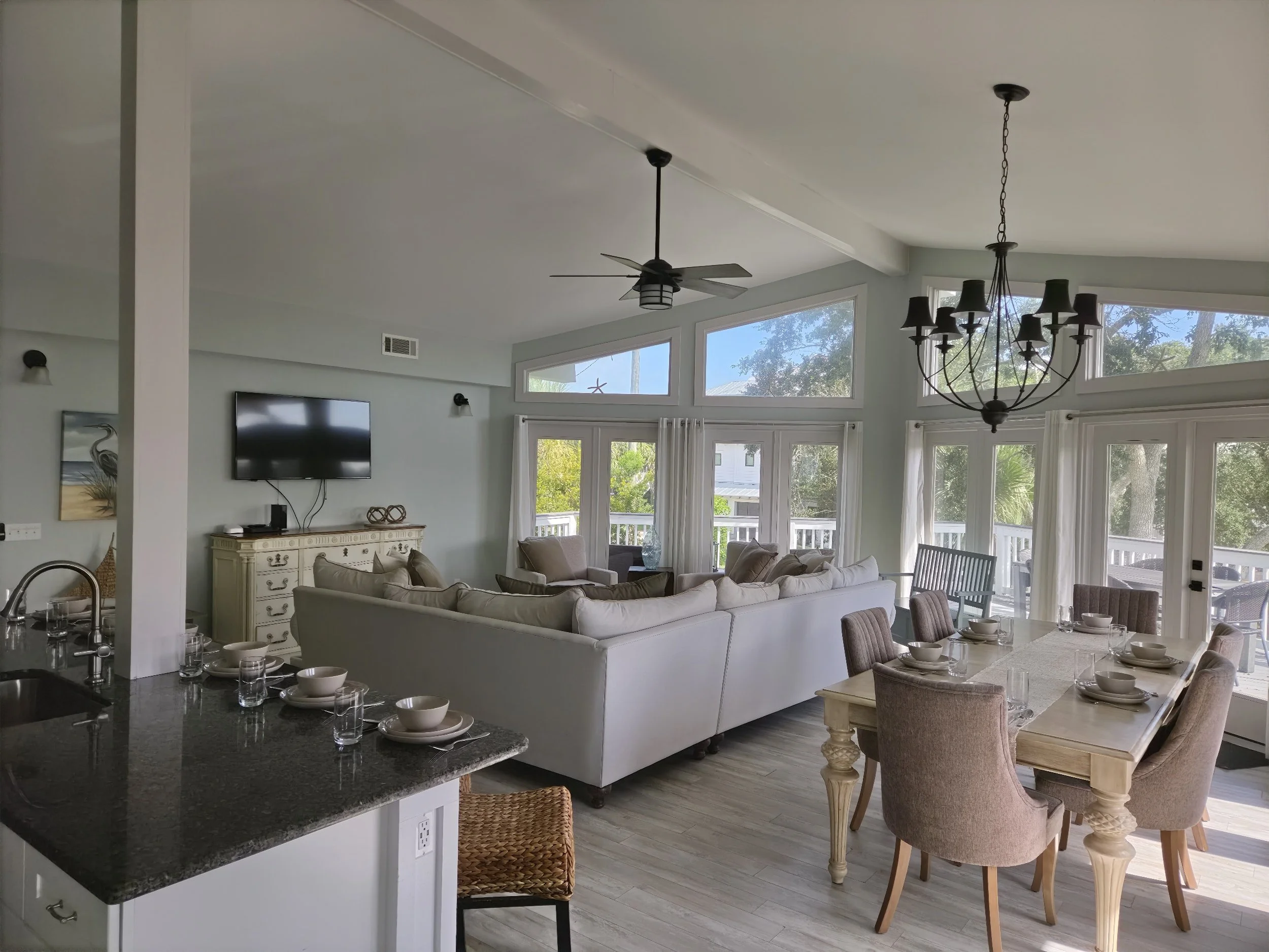 Bright open-concept living and dining room with a kitchen island, white sofas, ceiling fan, chandelier, television, and large windows overlooking a deck