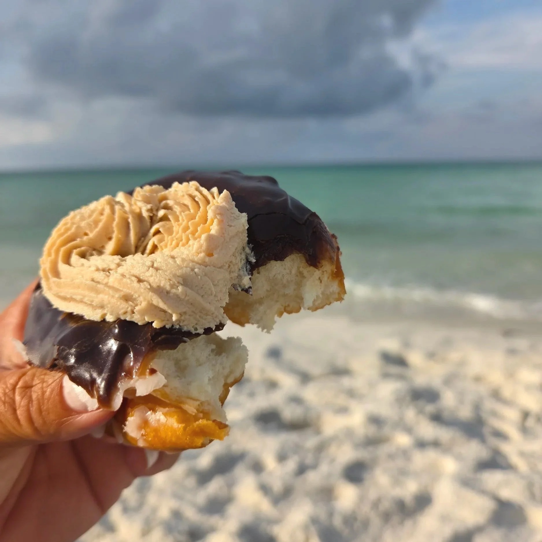 Hand holding a chocolate-covered ice cream sandwich on a beach with turquoise water in the background