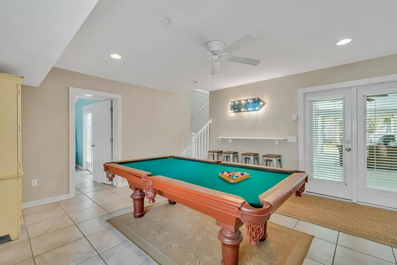 Bright beach house game room with a green pool table, colorful billiard balls, bar stools, ceiling fan, and illuminated beach house sign.