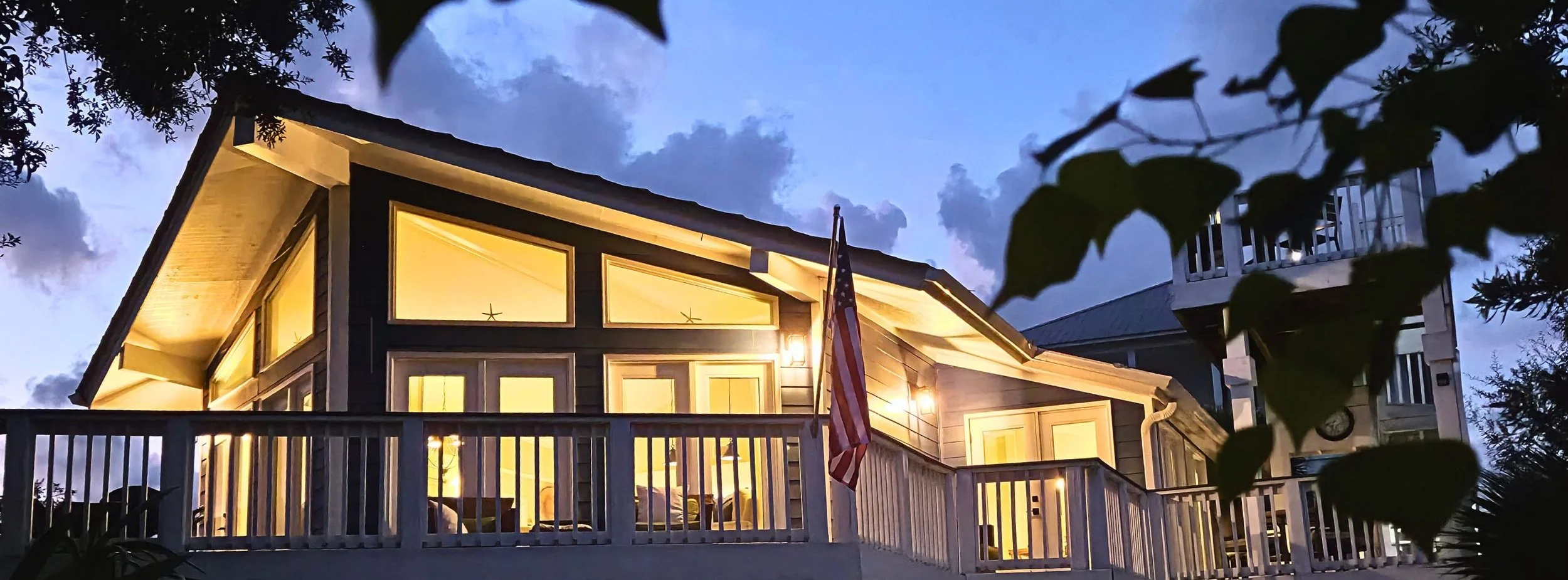 Two-story coastal house glowing with warm lights at dusk, featuring a porch, American flag, and surrounding foliage.