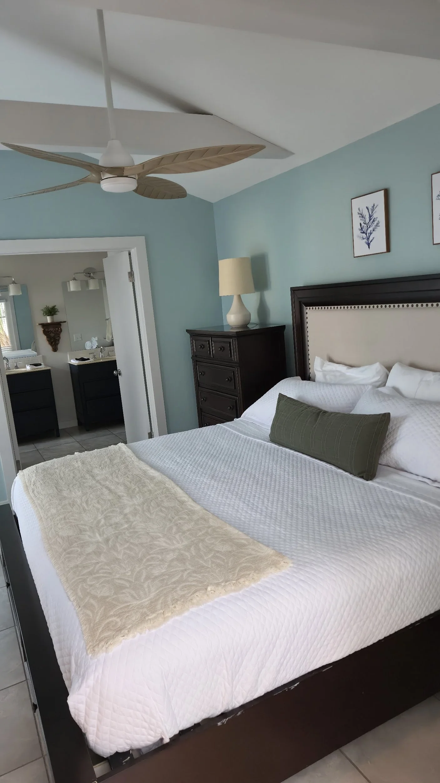 Bright bedroom with a neatly made bed, dark wood headboard and dresser, white bedding, ceiling fan, table lamp, and blue walls.