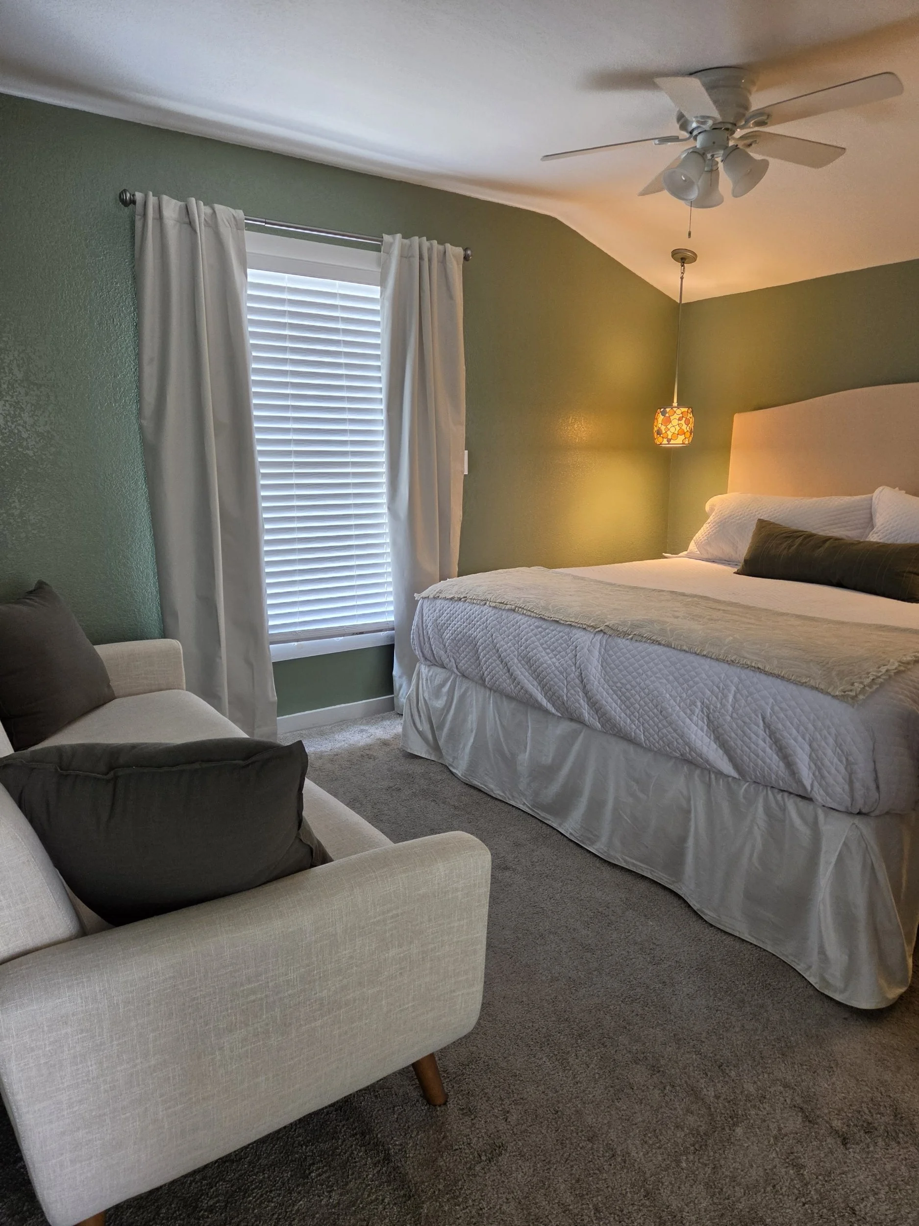 Cozy bedroom with a neatly made bed, green walls, white curtains, ceiling fan, pendant light, and upholstered armchair.