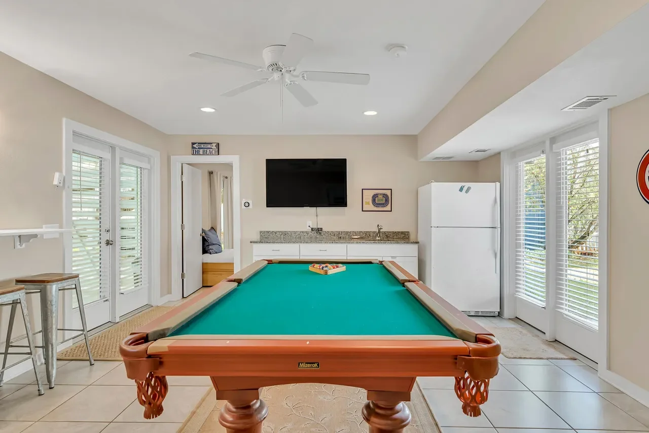 Bright game room with a green-felt pool table, wall-mounted TV, kitchenette, refrigerator, ceiling fan, and plantation-shuttered windows.