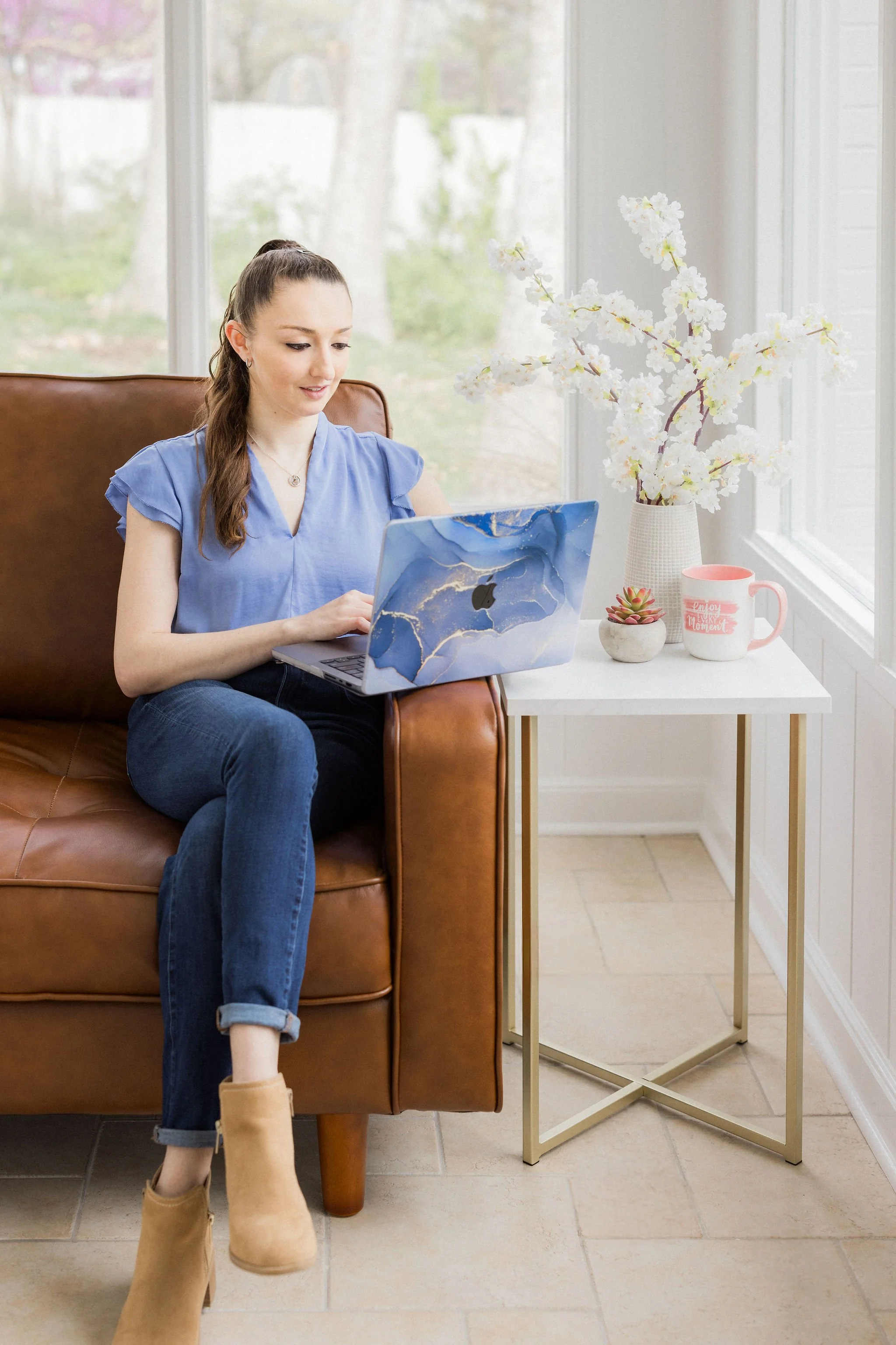A woman sitting on a leather chair next to a window, looking at her laptop, with a white flower vase and mug on a small table beside her.