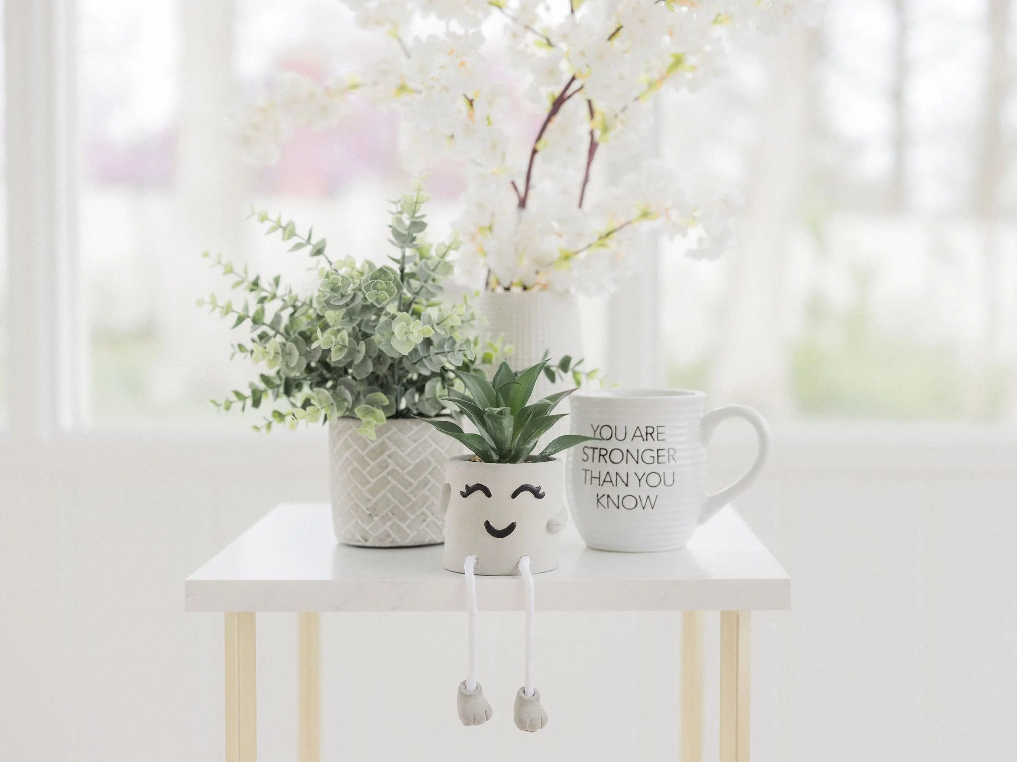 A white decorative mug with a smiling face, black eyebrows, and the text 'You are stronger than you know'. The mug has a rounded body and is filled with green and white plant stems and leaves. It is placed on a white stand against a blurry background of light-colored curtains and window.
