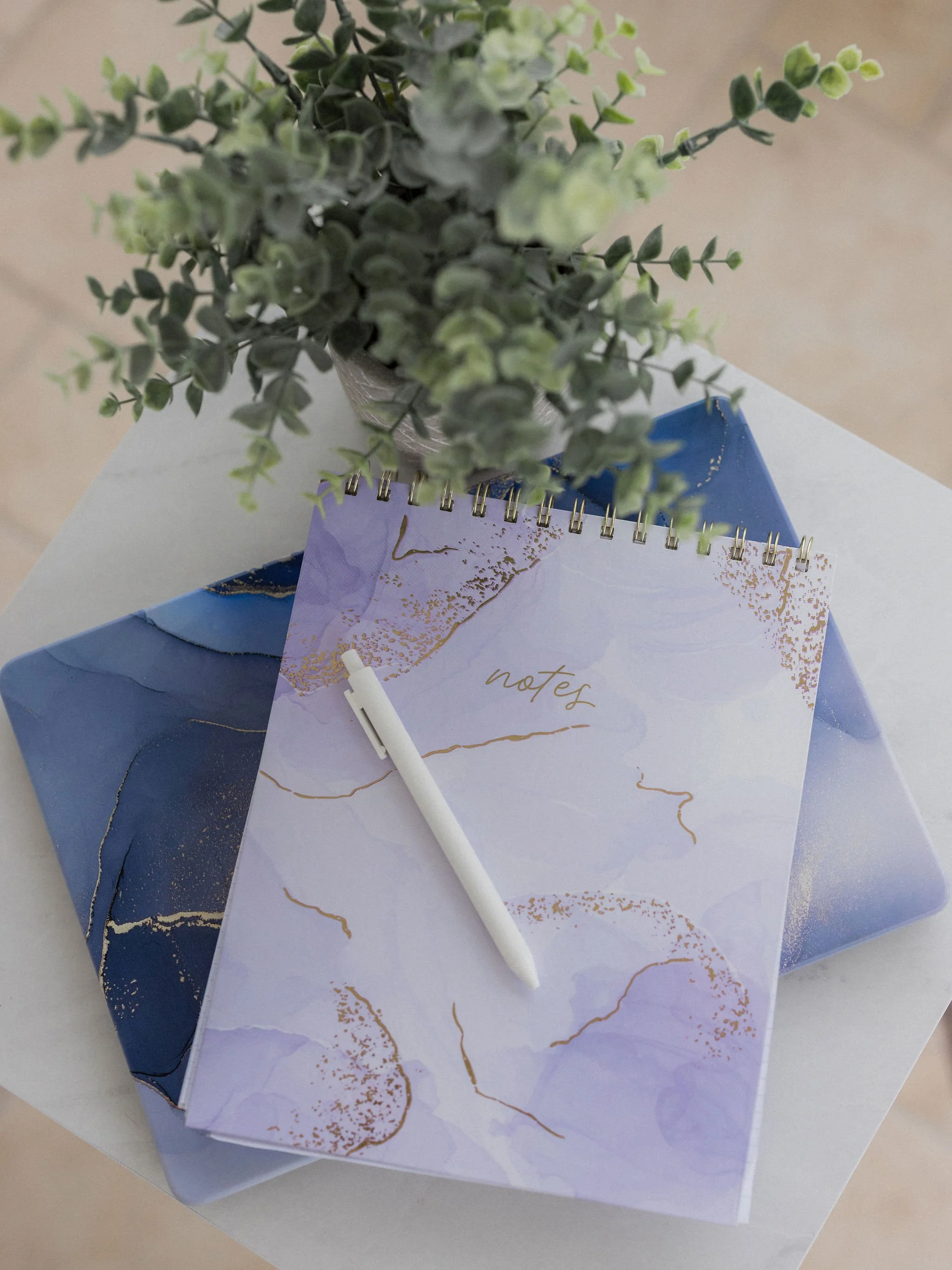 A potted artificial green plant placed on top of a small notebook with a pen resting on it. The table has a decorative blue cloth with gold accents. There are also two sheets of marbled paper in pastel purple and blue colors underneath the plant and notebook.