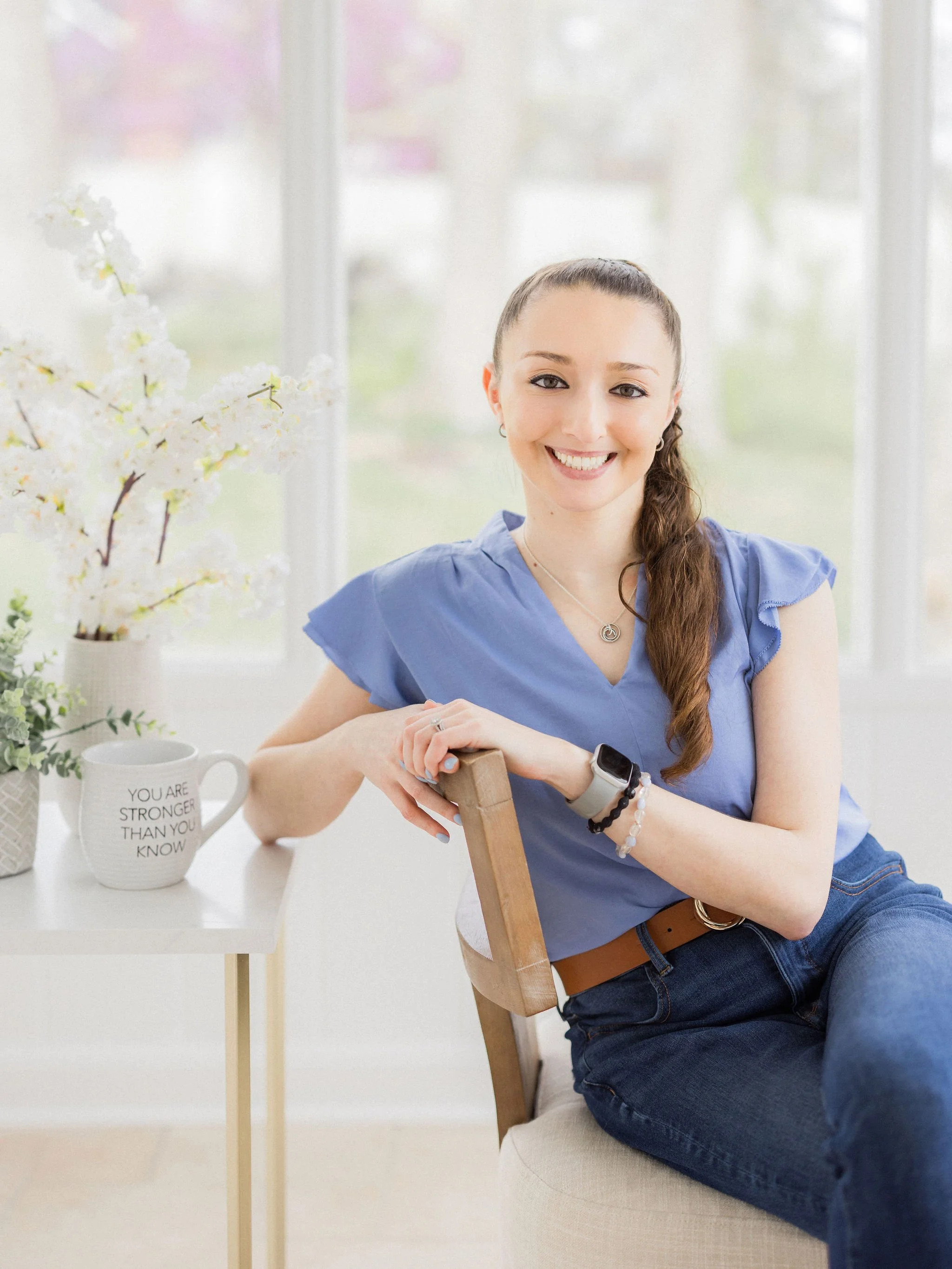 A woman with long brown hair in a braid, wearing a blue top, jeans, and a smartwatch, is sitting on a beige chair at a white table, smiling at the camera. There is a potted flowering plant and a white mug with motivational text on the table and a bright, airy room with white curtains in the background.