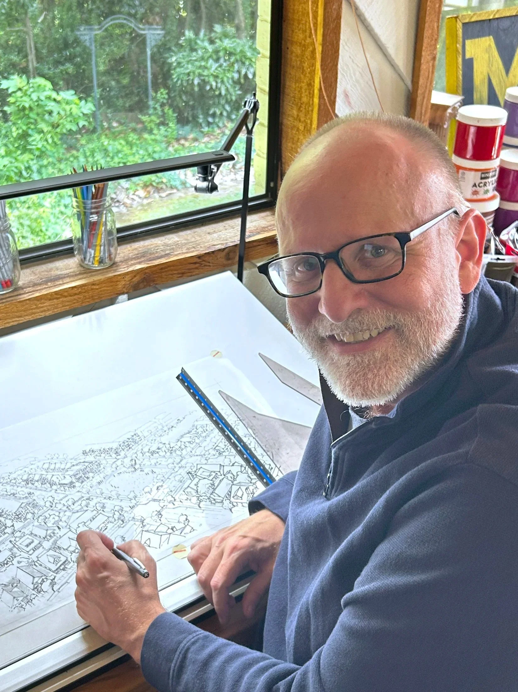 Portrait of a designer drawing or planning on an architectural blueprint or map at a desk, with a window showing greenery outside behind him.