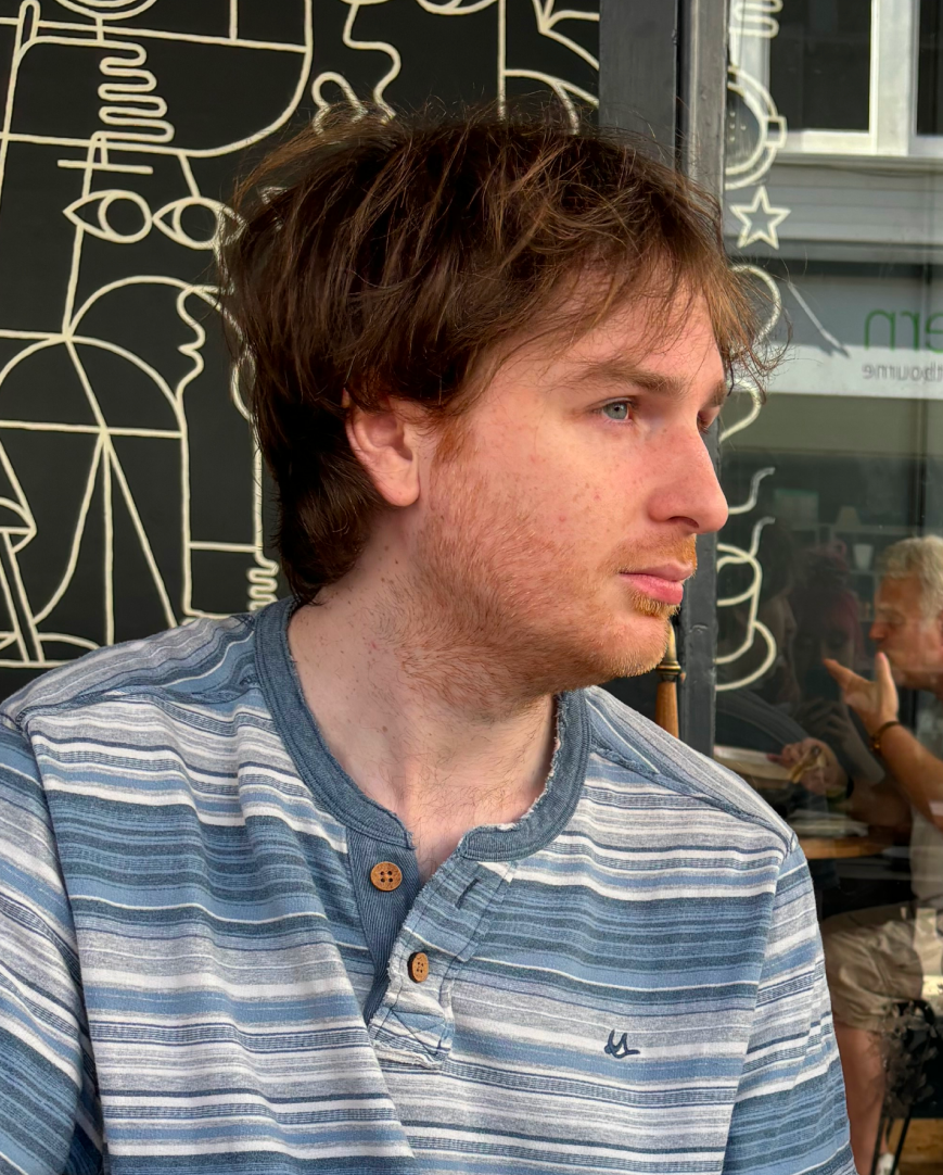 A young man with medium-length brown hair and light facial hair, wearing a blue and gray striped shirt, sitting at an outdoor cafe. Behind him, there is a black wall with white abstract line art and a window reflecting people sitting inside.