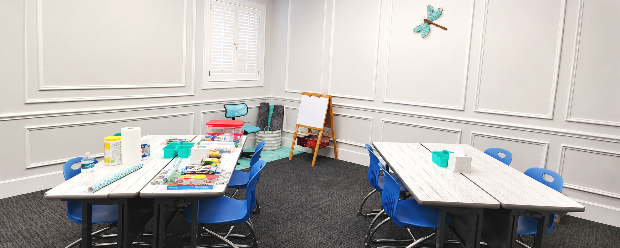 A room with white paneled walls set up for a children's activity or learning session, featuring two white tables surrounded by blue chairs, art supplies, tissue box, water bottles, and small storage containers, with a decorative blue dragonfly wall art.