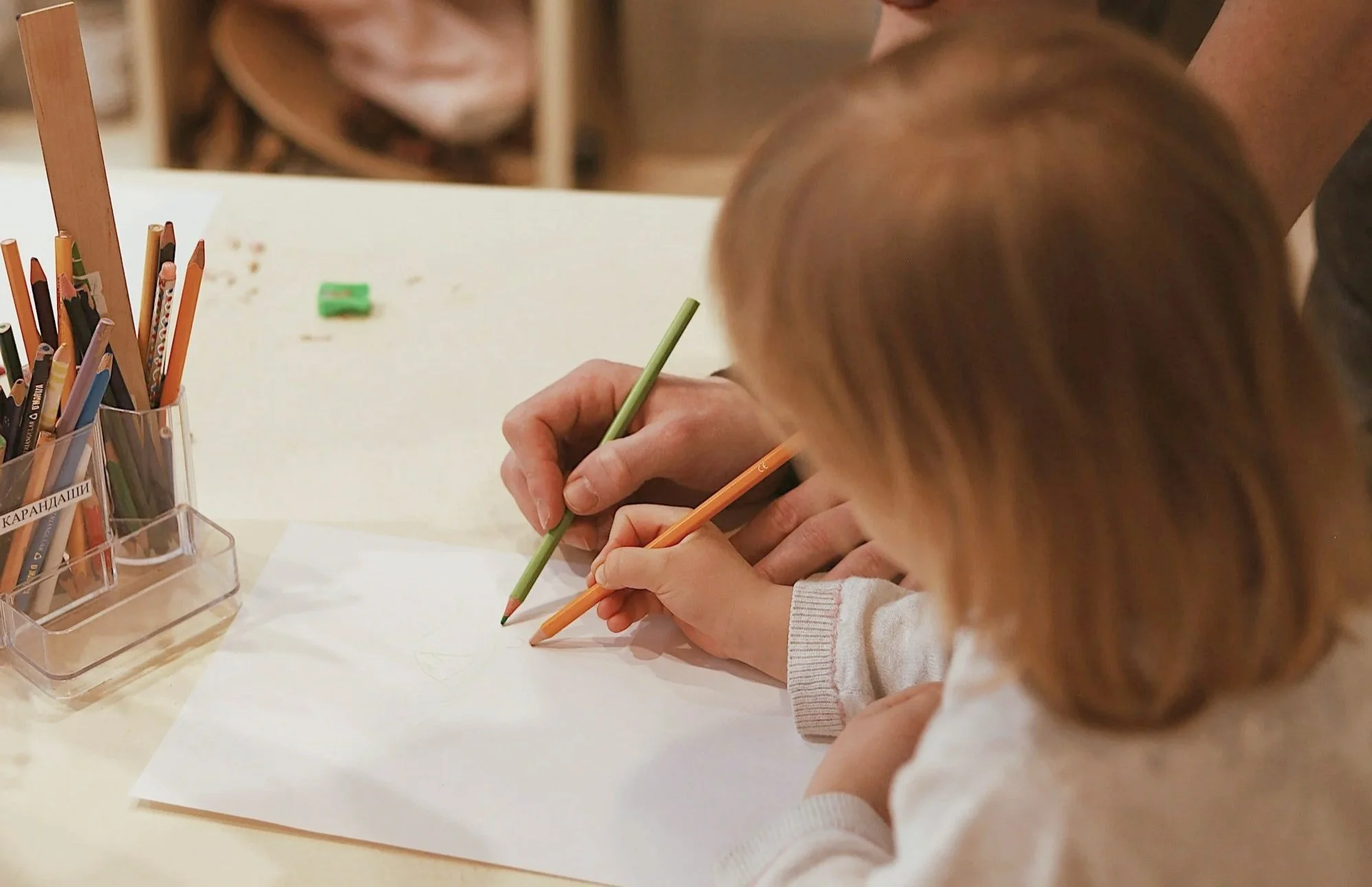 Two children drawing with colored pencils on white paper, with a container of colored pencils on the table in front of them.