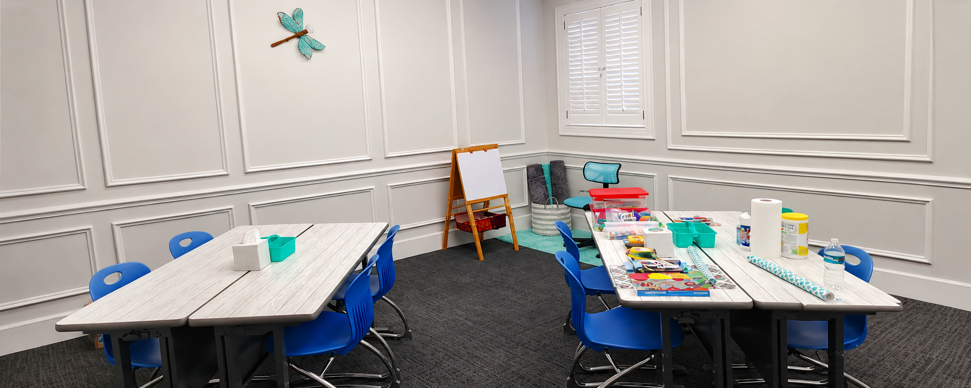A children's craft room with two white tables, blue chairs, and various craft supplies like paper, markers, and tape. The walls are decorated with a blue dragonfly wall hanging, and there is a window with white shutters. The room has white paneled walls and dark carpet flooring.