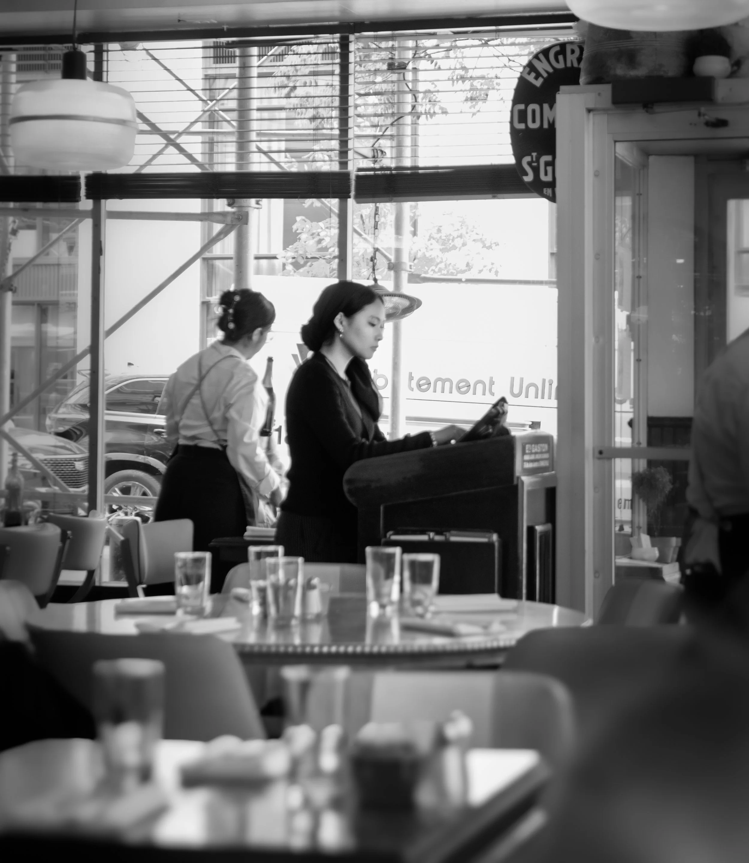 Two women, one with dark hair and the other with light hair, are standing and possibly working or talking in a modern indoor space with large windows and visible outdoor scaffolding.