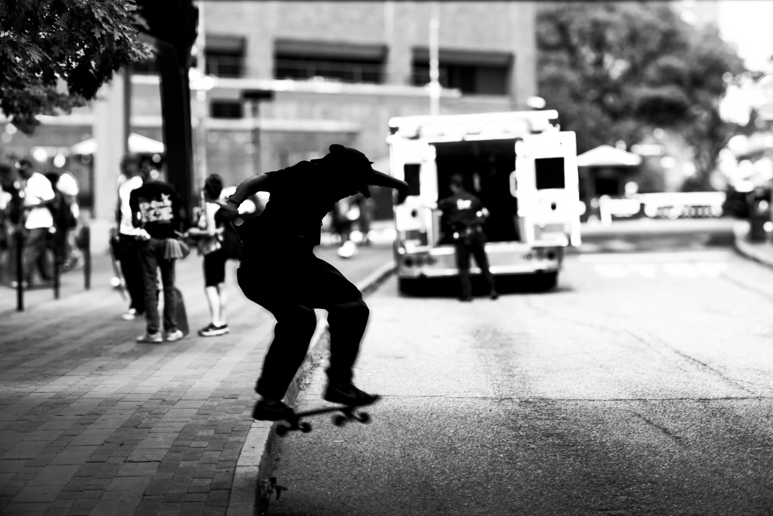A person skateboarding on a city sidewalk, with several pedestrians in the background near buildings and trees, all in black and white.