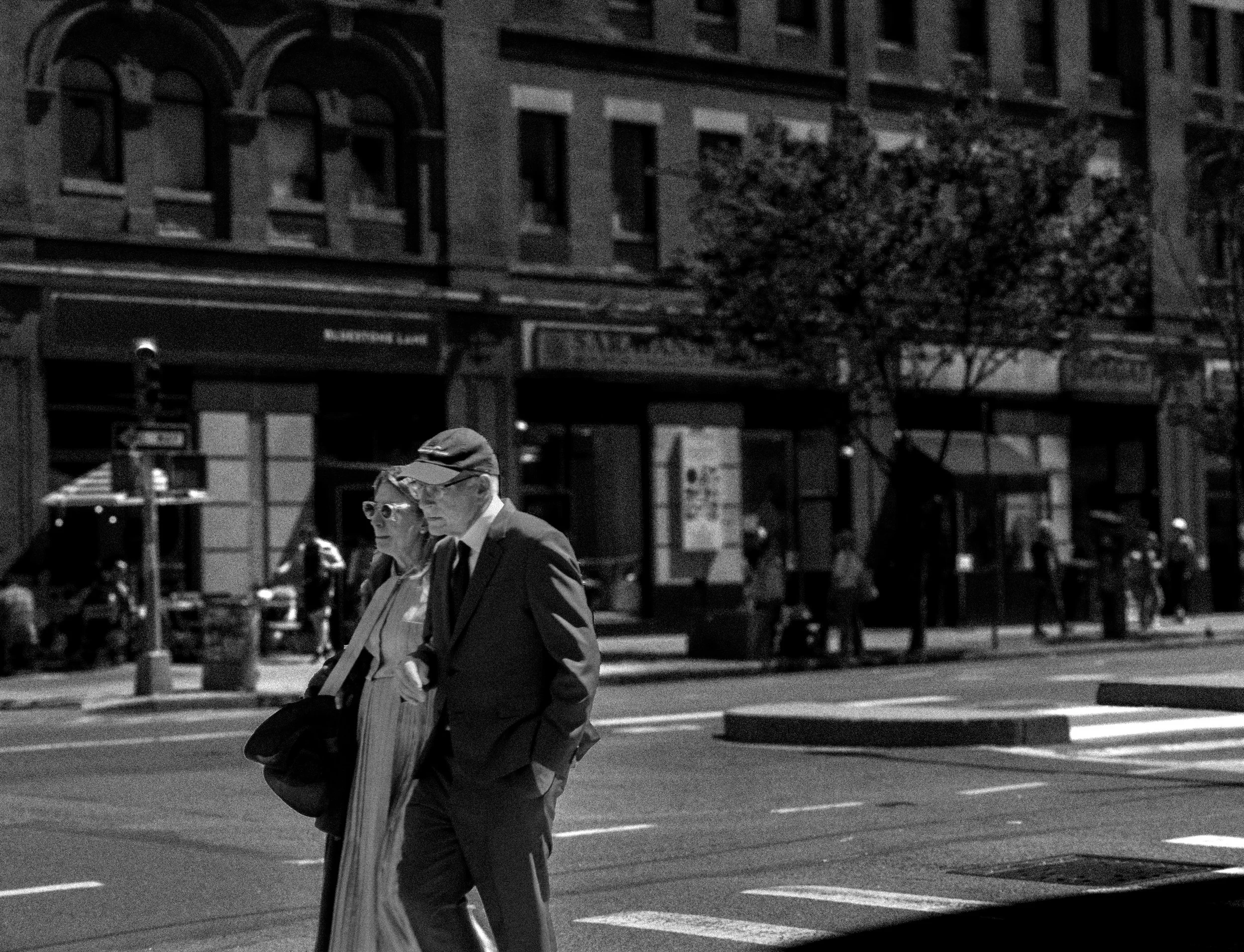 Two elderly people, a man and a woman, walking on a city street at night. The man is wearing glasses, a cap, and a jacket, while the woman is wearing sunglasses, earrings, and a coat. They are walking past buildings with lit windows and storefronts.