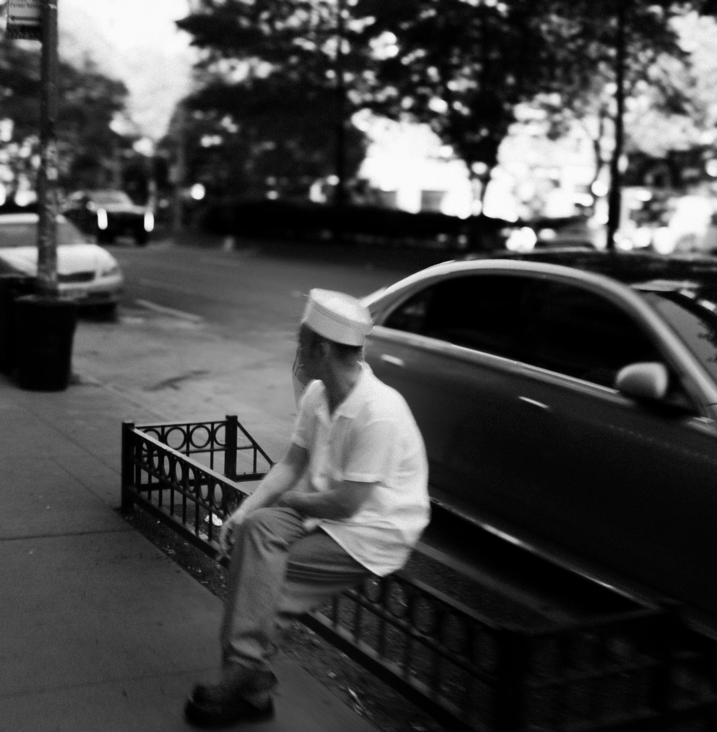 Black and white photo of a person walking on a sidewalk with trees and parked cars in the background. The person is seen from behind, wearing a light-colored shirt and dark pants, holding a bag, and walking past a bicycle rack.
