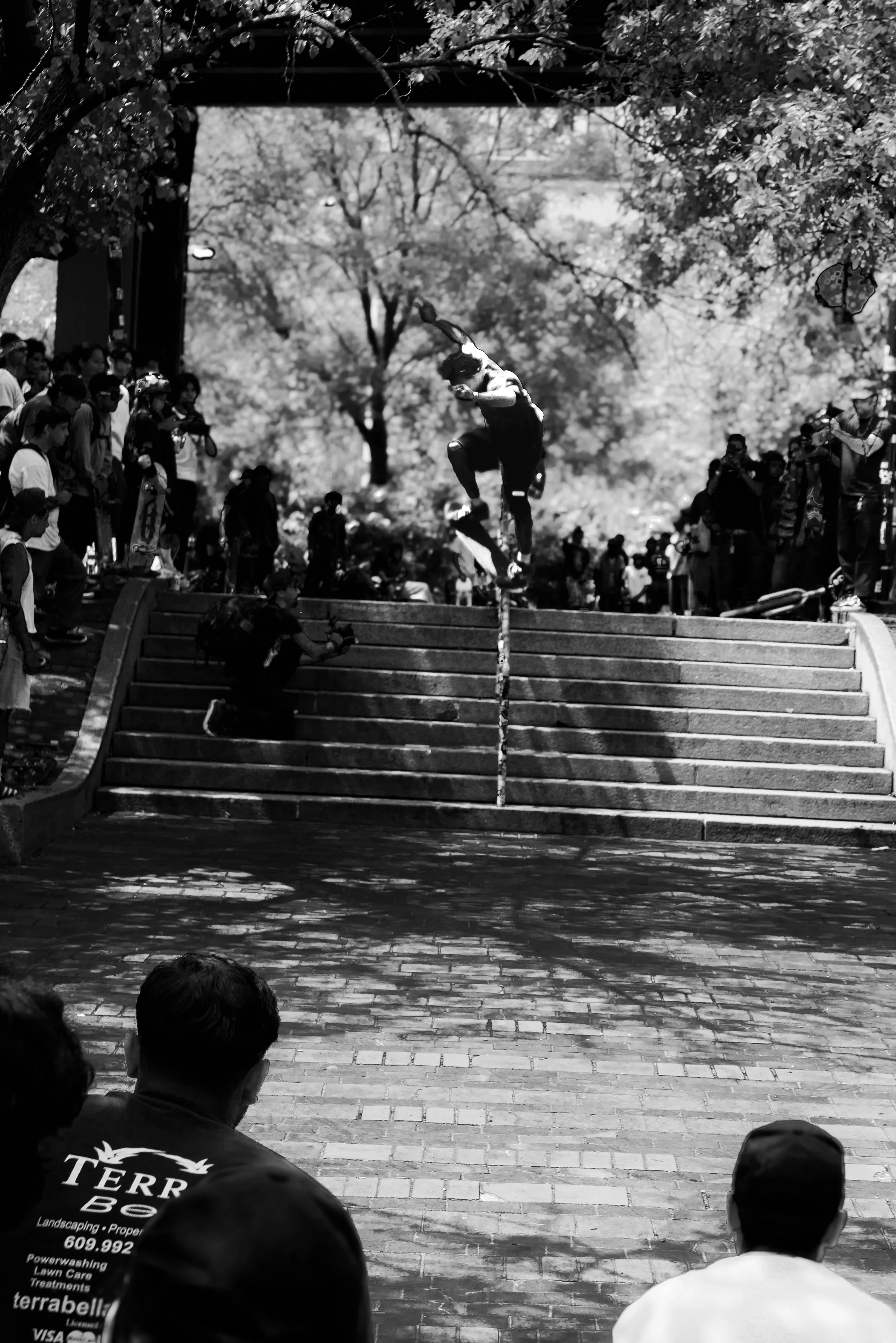 A black and white photo of a crowd of people watching a skateboarder perform a trick on stairs in a park with trees in the background.
