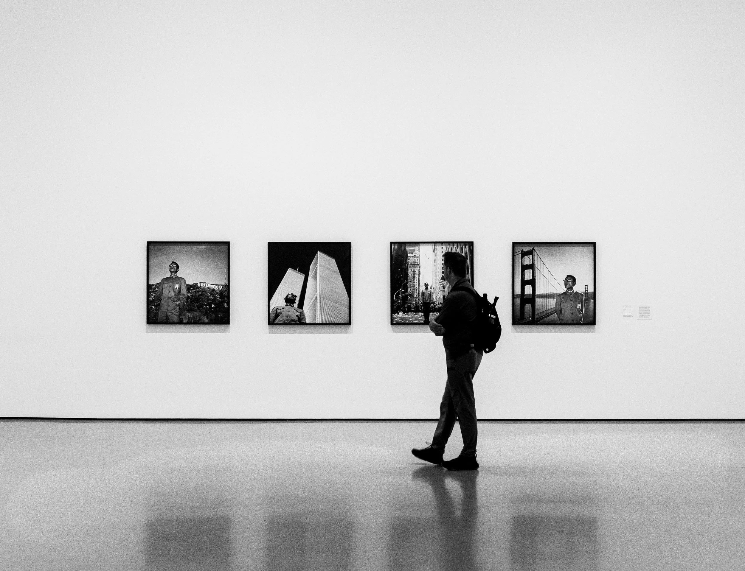A person with a backpack walking in front of four framed photographs displayed on a white gallery wall.
