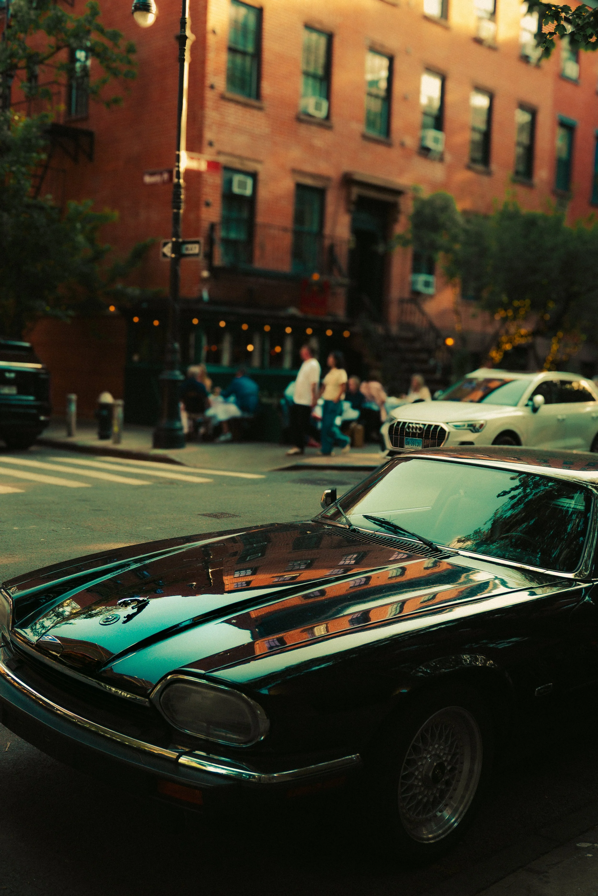 A parked black vintage car with a sleek, rounded design and a shiny exterior reflecting the buildings around it, in front of a city street scene with a building facade, people sitting at an outdoor cafe, and a street sign.