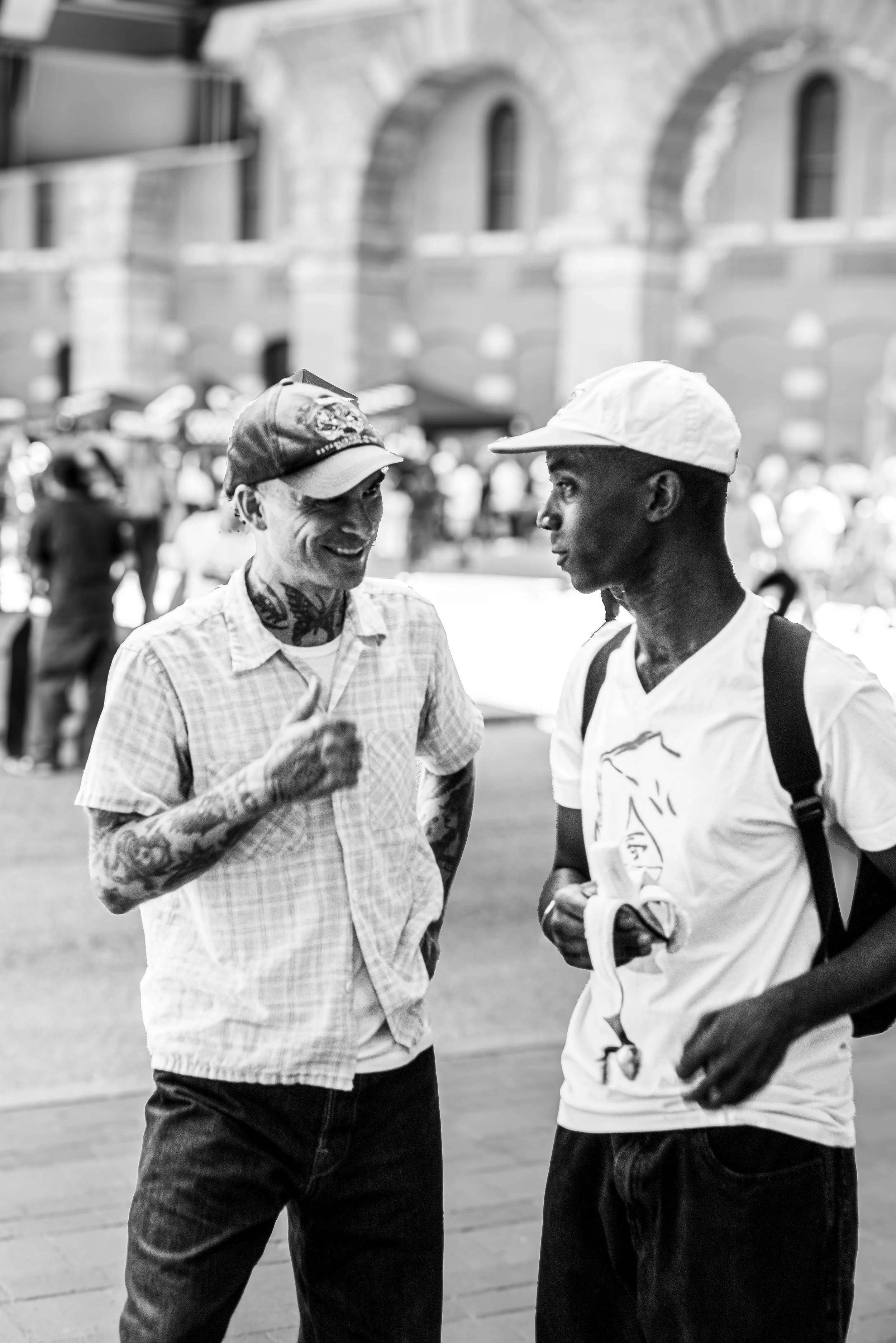 Two men having a conversation on a city street, one smiling and the other listening, with a historic building in the background, in black and white.