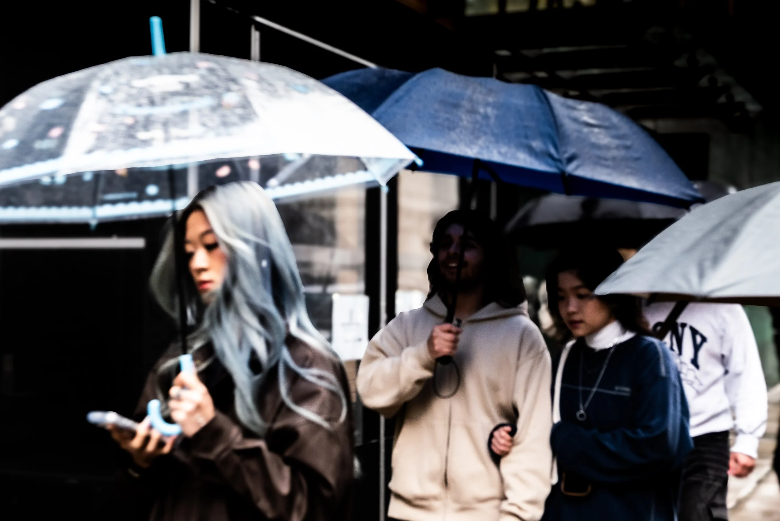 Group of people walking outdoors in rainy weather, holding umbrellas, with one person looking at a phone.