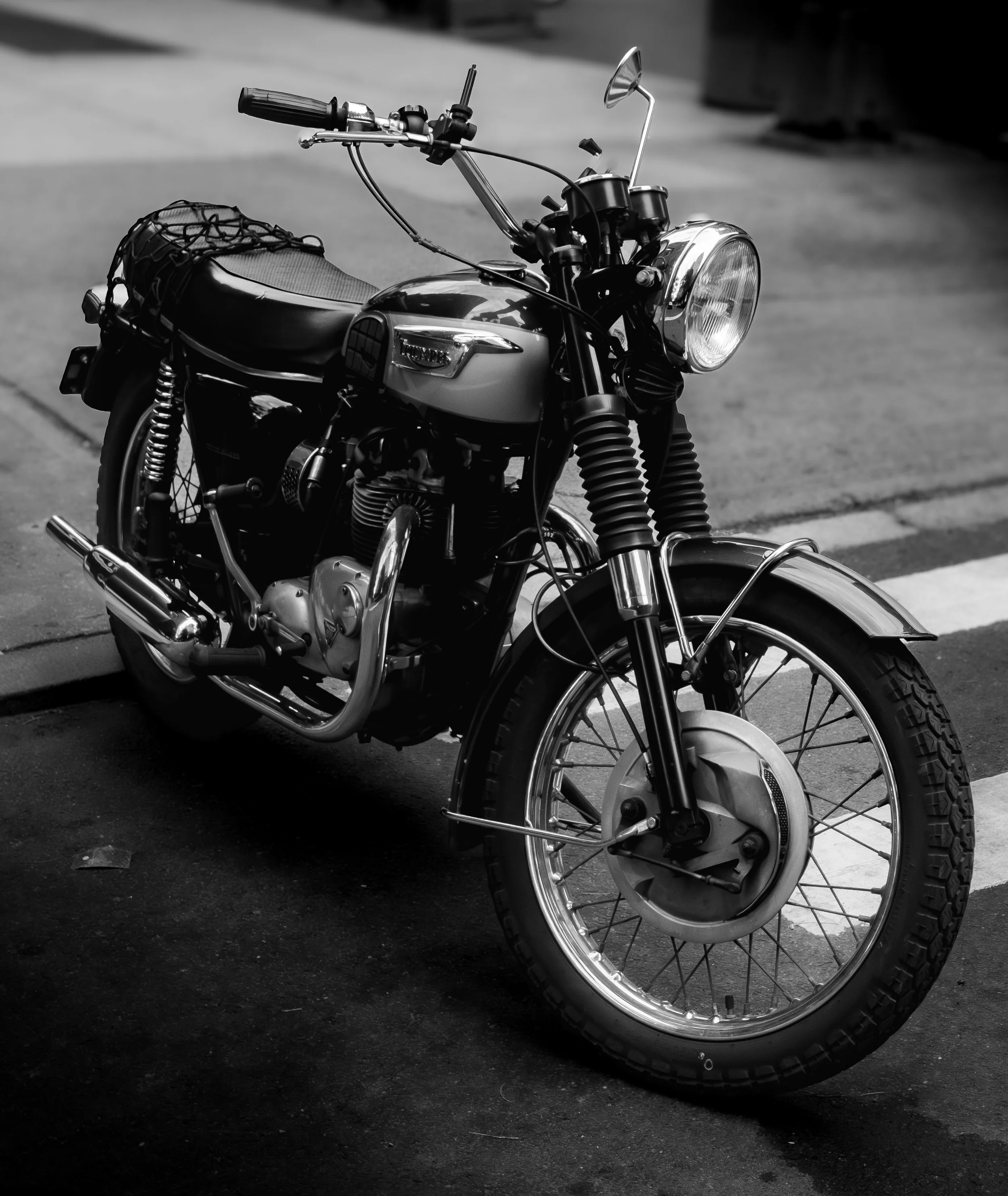 Black and white photograph of two motorcycles parked on a street, with one in the foreground and the other slightly behind, showing various parts including tires, handlebars, and mirrors.