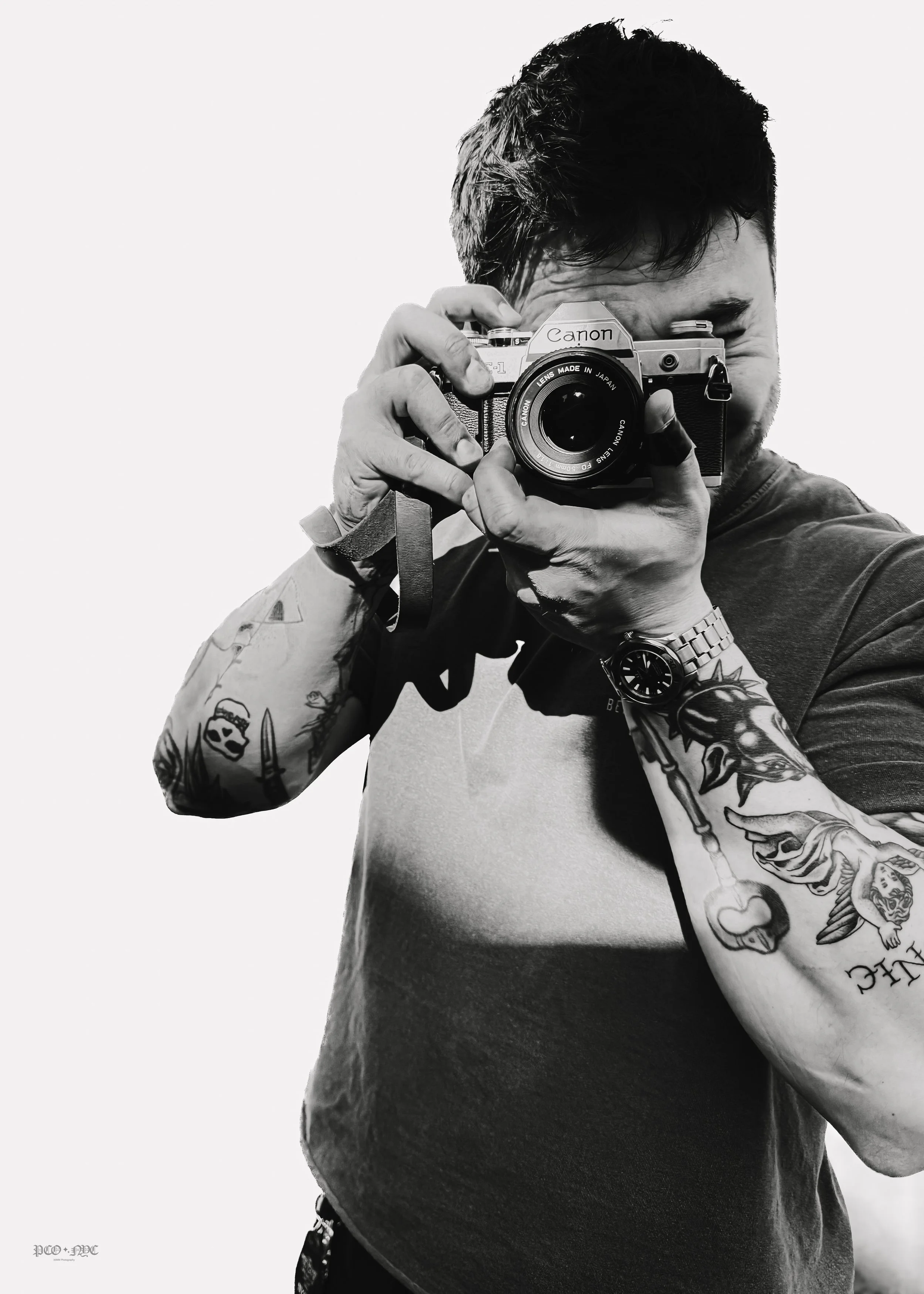 A man with tattoos on his arms and wearing a wristwatch is taking a photo of himself with a vintage camera. The photo is black and white, and the man's face is partly covered by the camera.